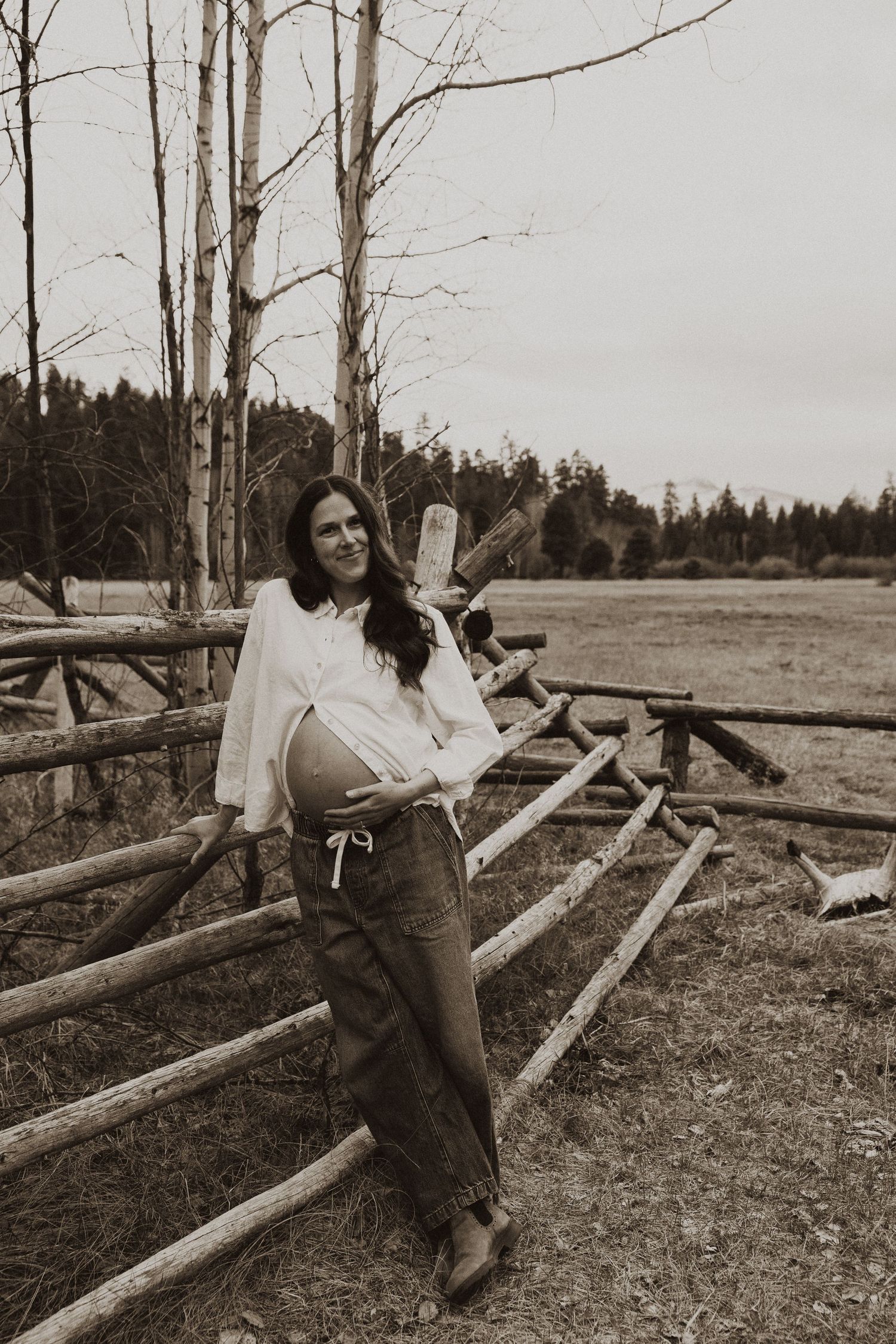 Sepia photo of pregnant woman leaning against rustic wooden fence in white shirt and olive pants.