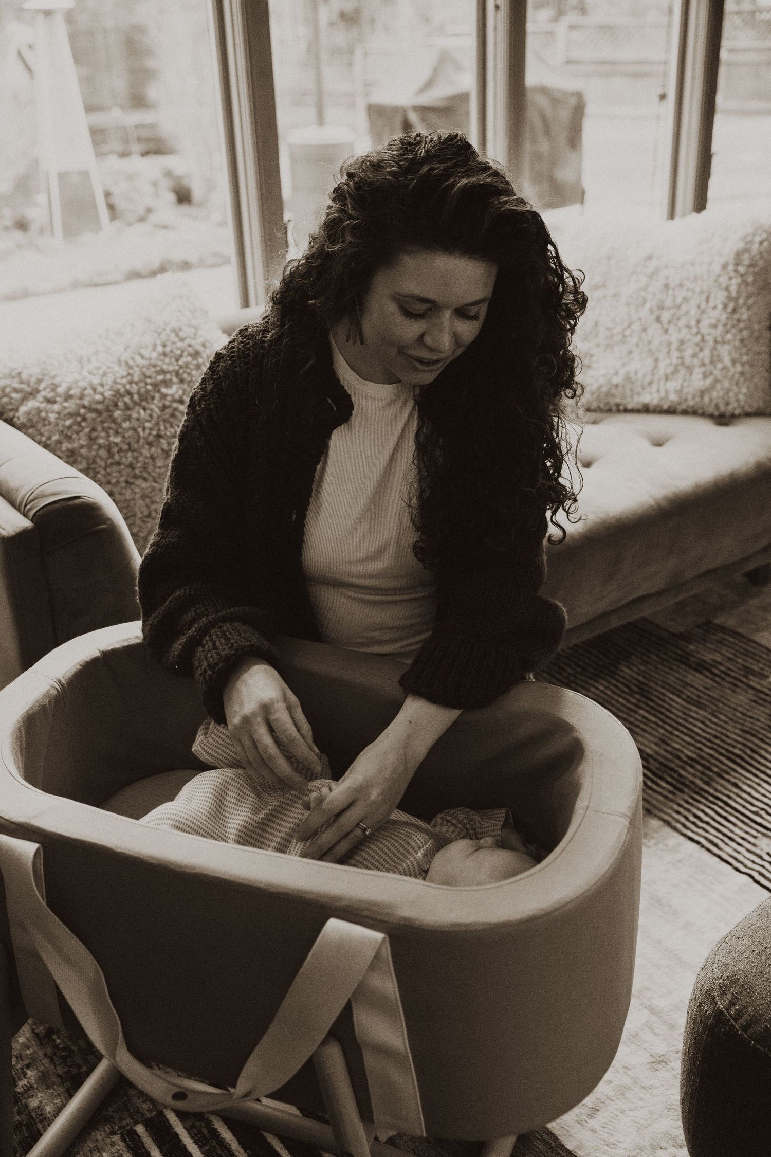 Curly-haired mother gently tending to her newborn baby in a mustard yellow bassinet.