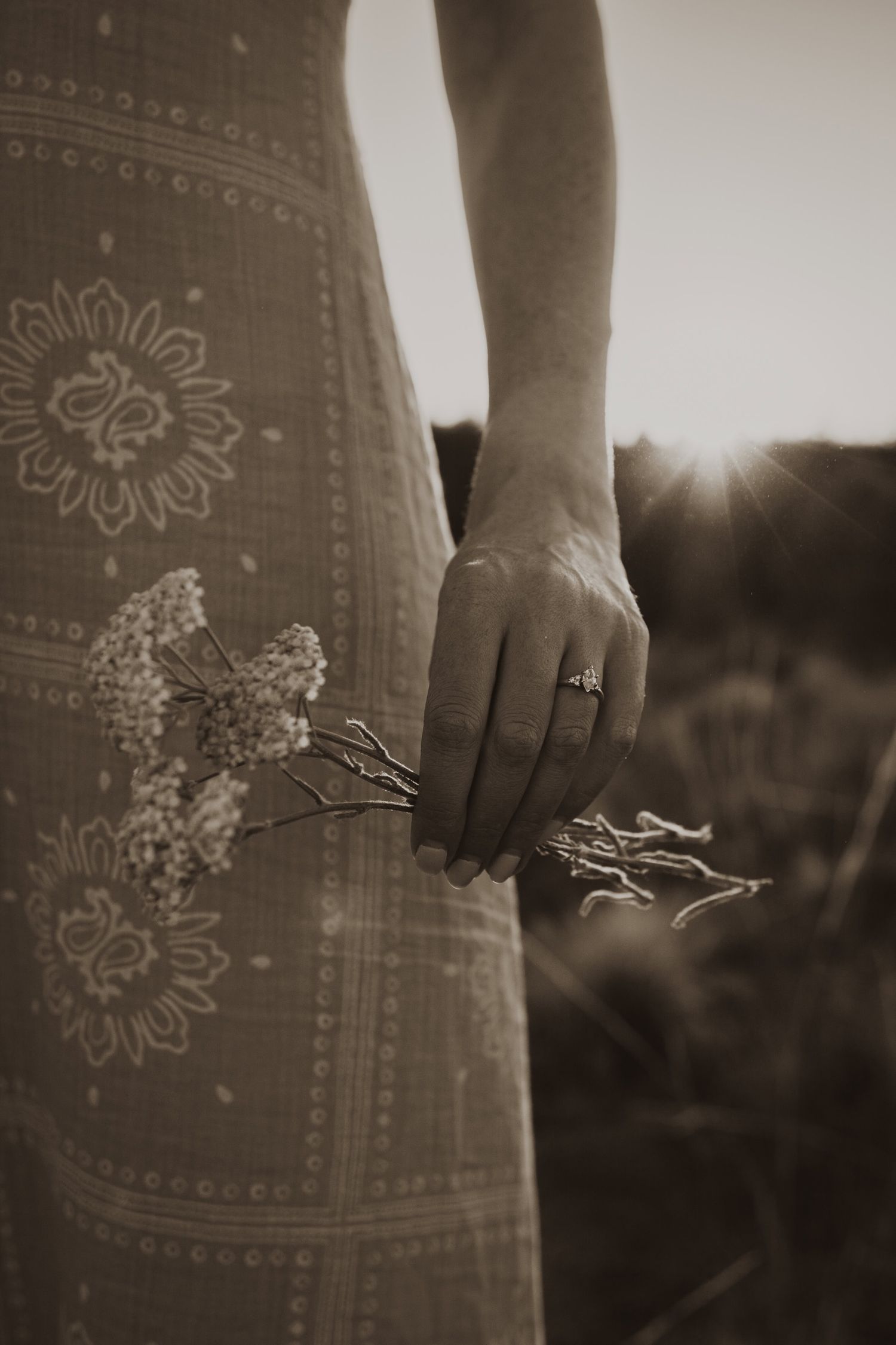 A close-up of a hand wearing an engagement ring against a patterned dress in golden sunlight.