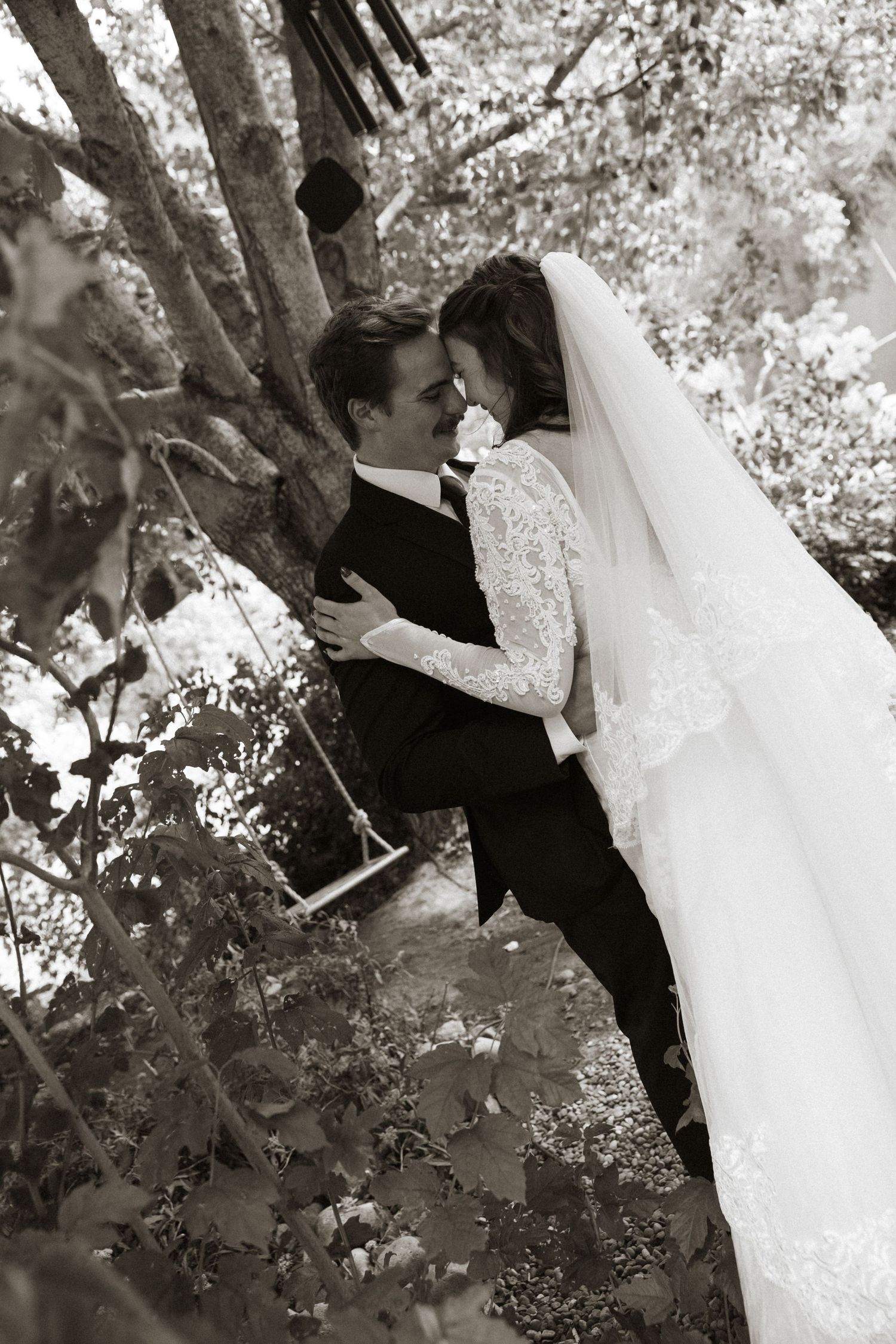 Black and white photo of bride and groom embracing near a tree with a swing in a garden setting.