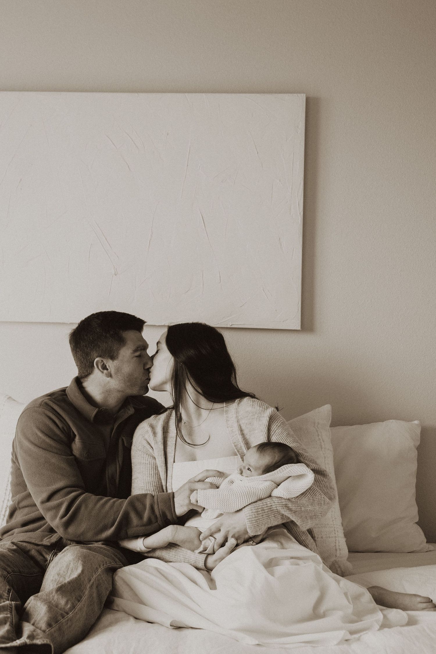 A couple shares a tender kiss while holding their newborn baby on a bed in a black and white photo.