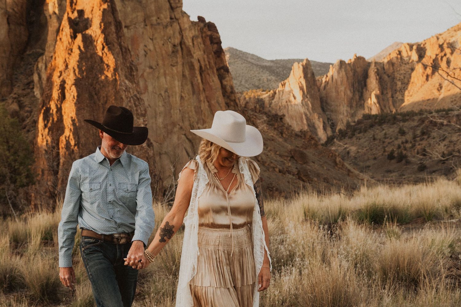Couple in western hats holding hands walking through a desert canyon with golden rock formations at sunset.