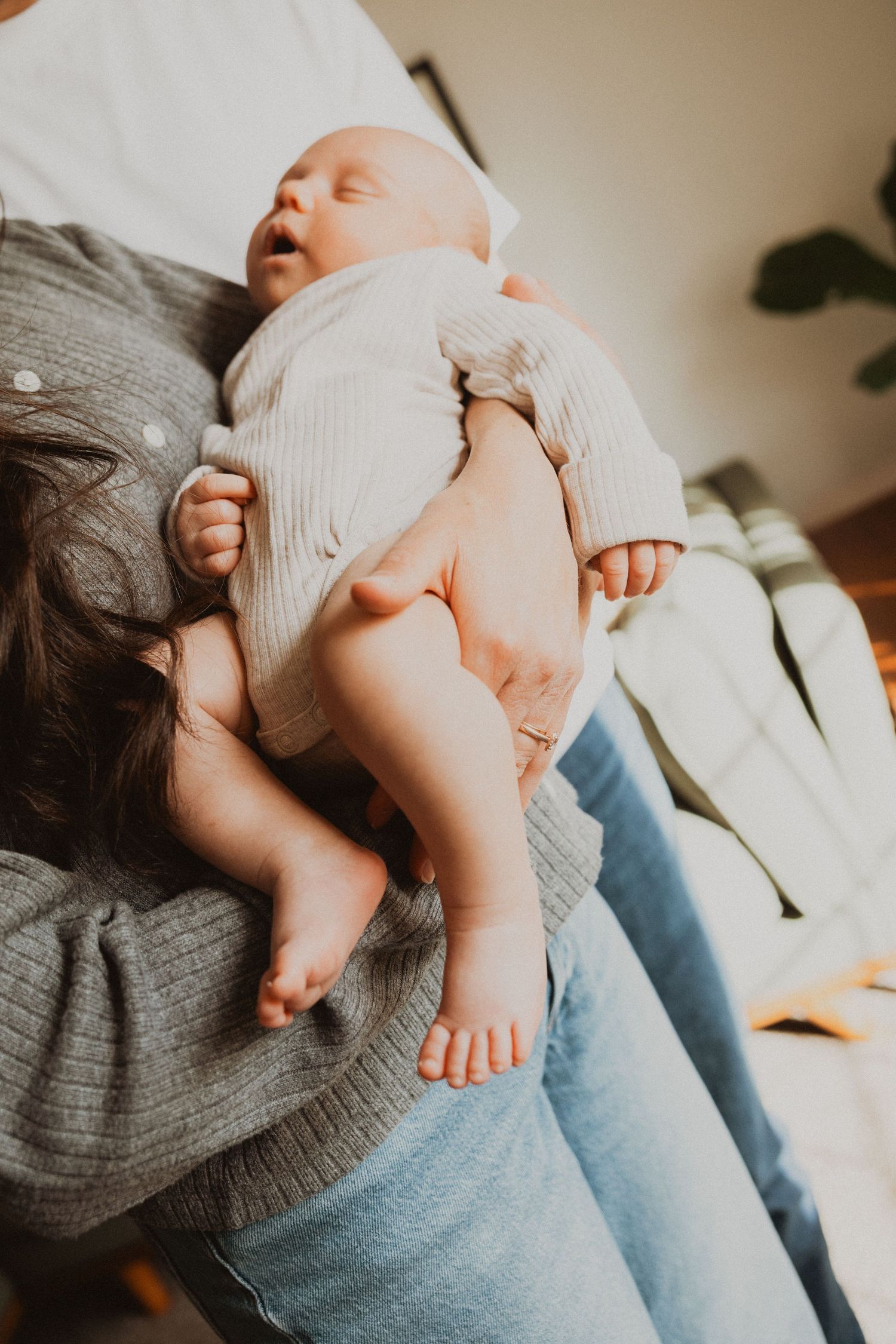 Mother cradling a sleeping newborn baby in her arms, showing tiny bare feet in soft natural light.