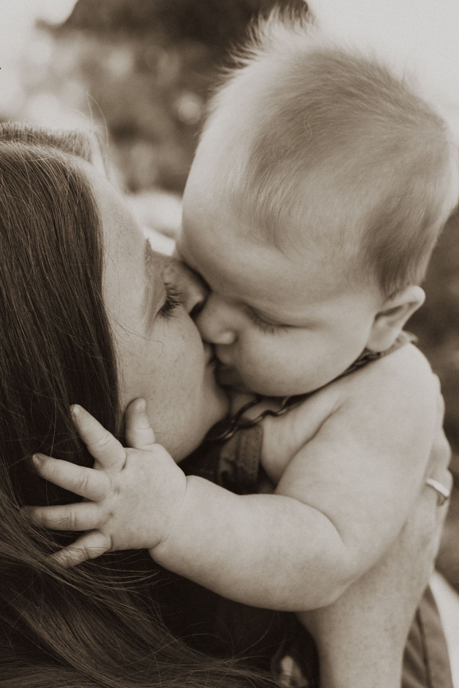 A tender sepia-toned close-up of a mother and baby sharing a sweet nose-to-nose kiss outdoors.