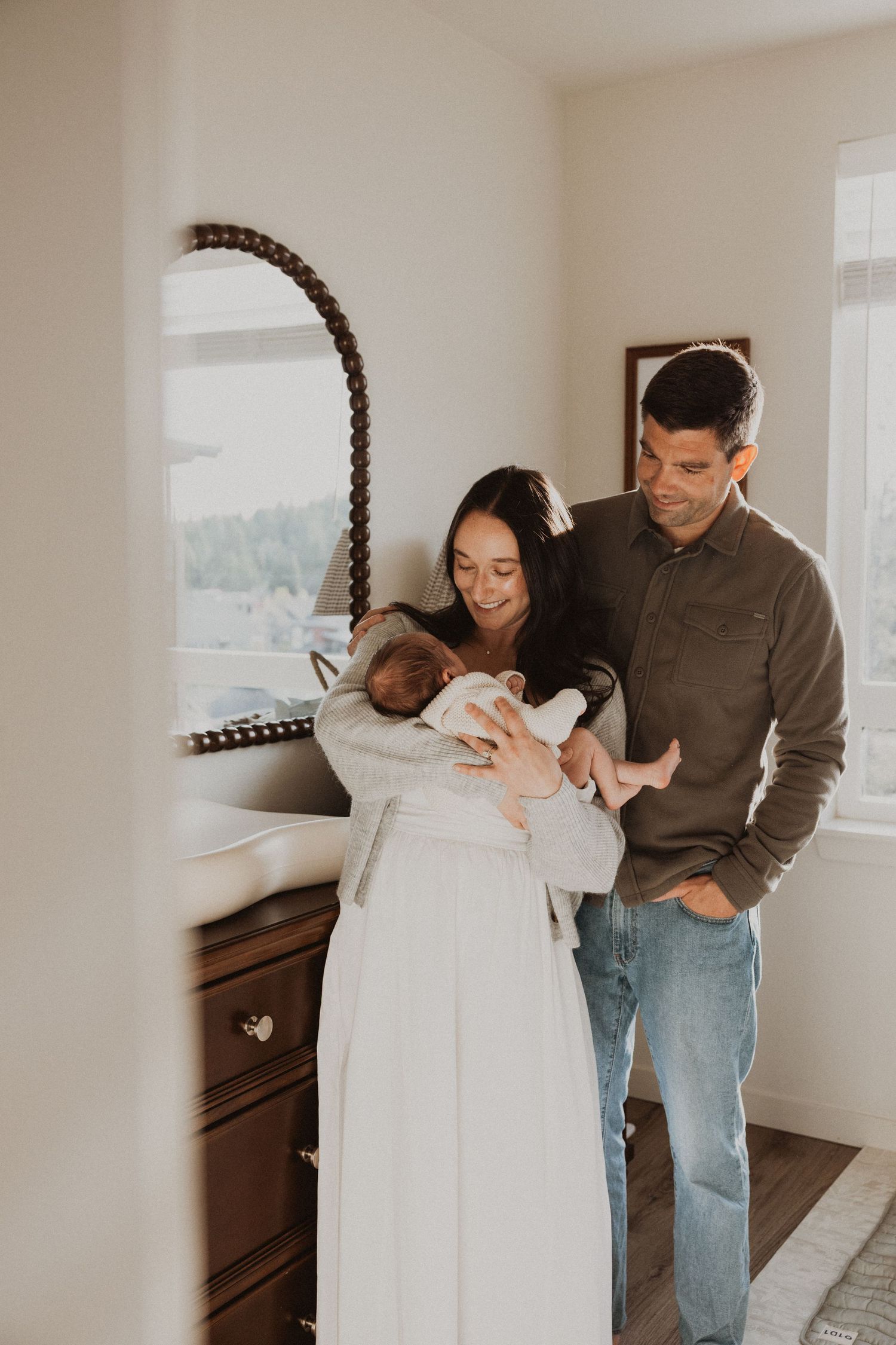 A couple smiles lovingly at their newborn baby in a bright, cozy bedroom with wooden furniture.