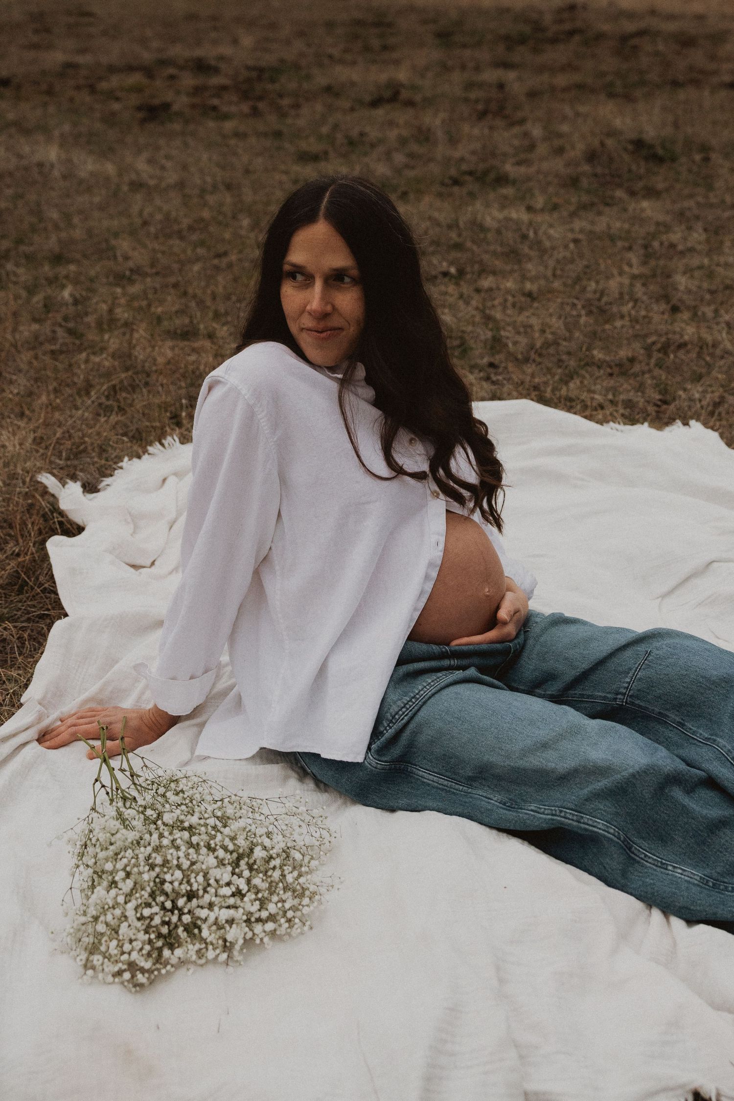 Pregnant woman in white blouse reclines on blanket holding baby's breath bouquet in an open field.