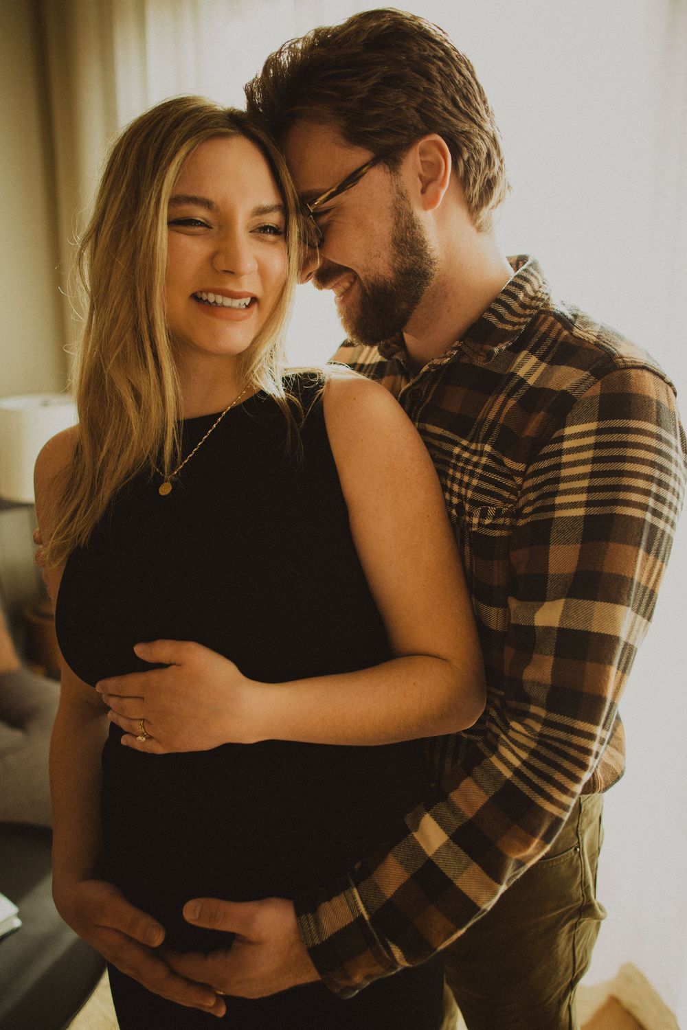 Intimate couple embracing and smiling in warm sunlight by a window wearing casual attire.