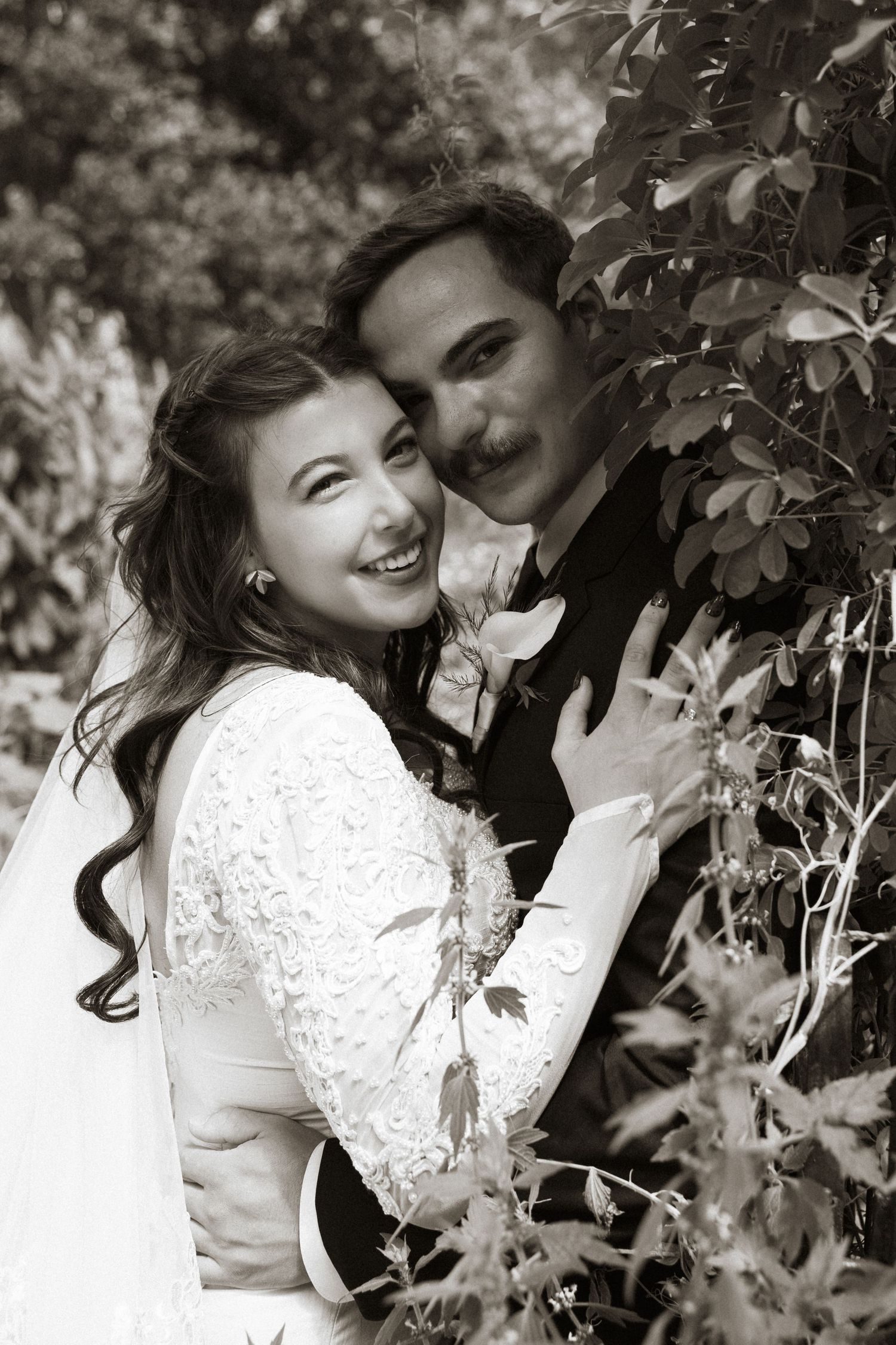 Black and white portrait of smiling bride in lace gown embracing groom among leafy vines outdoors.