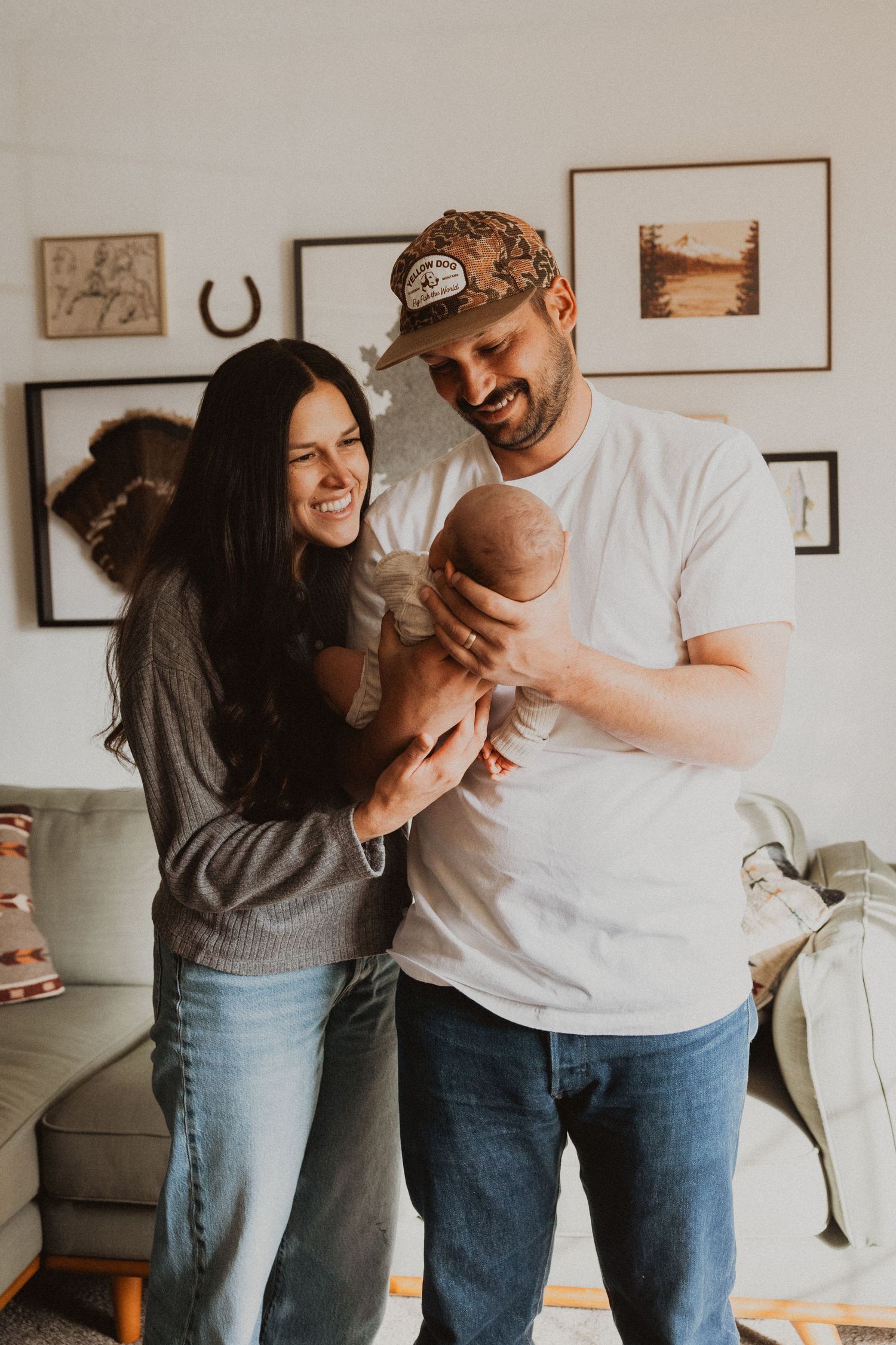 Happy new parents smiling and holding their newborn baby in a cozy living room with framed art on the wall.