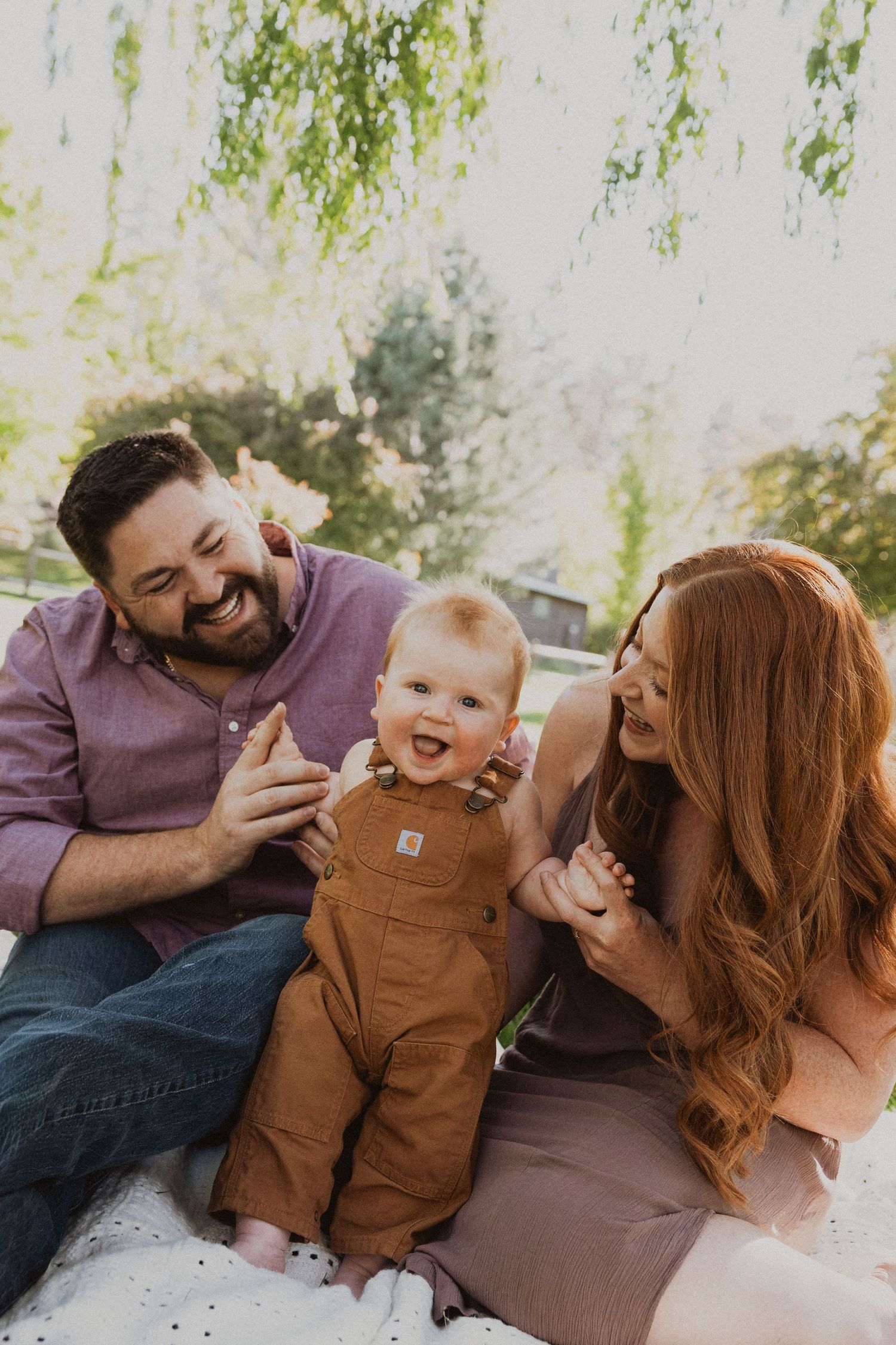Happy family with laughing baby in brown overalls sitting outdoors with green trees in background.