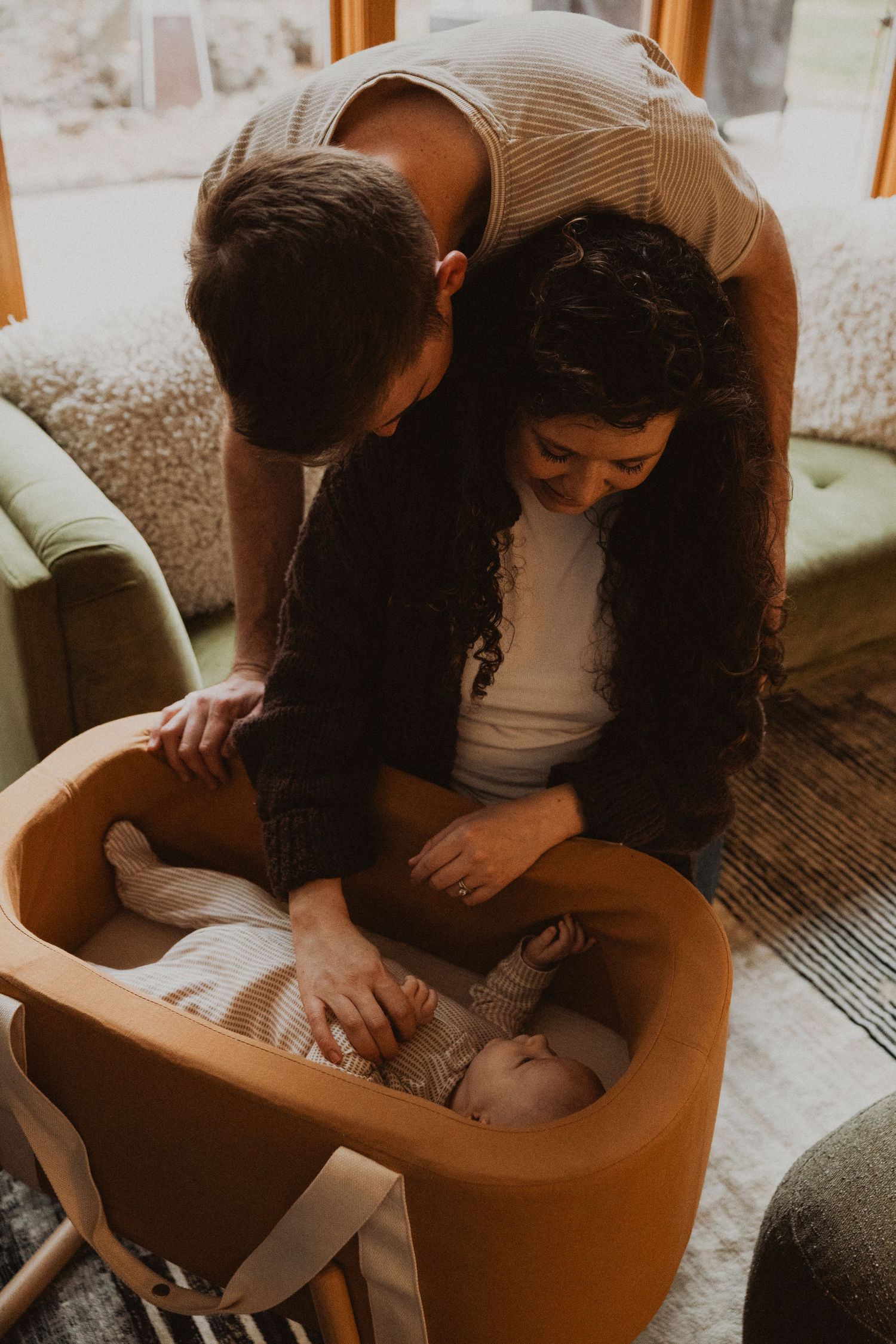 New parents lovingly gazing at their newborn baby resting in a mustard yellow bassinet.