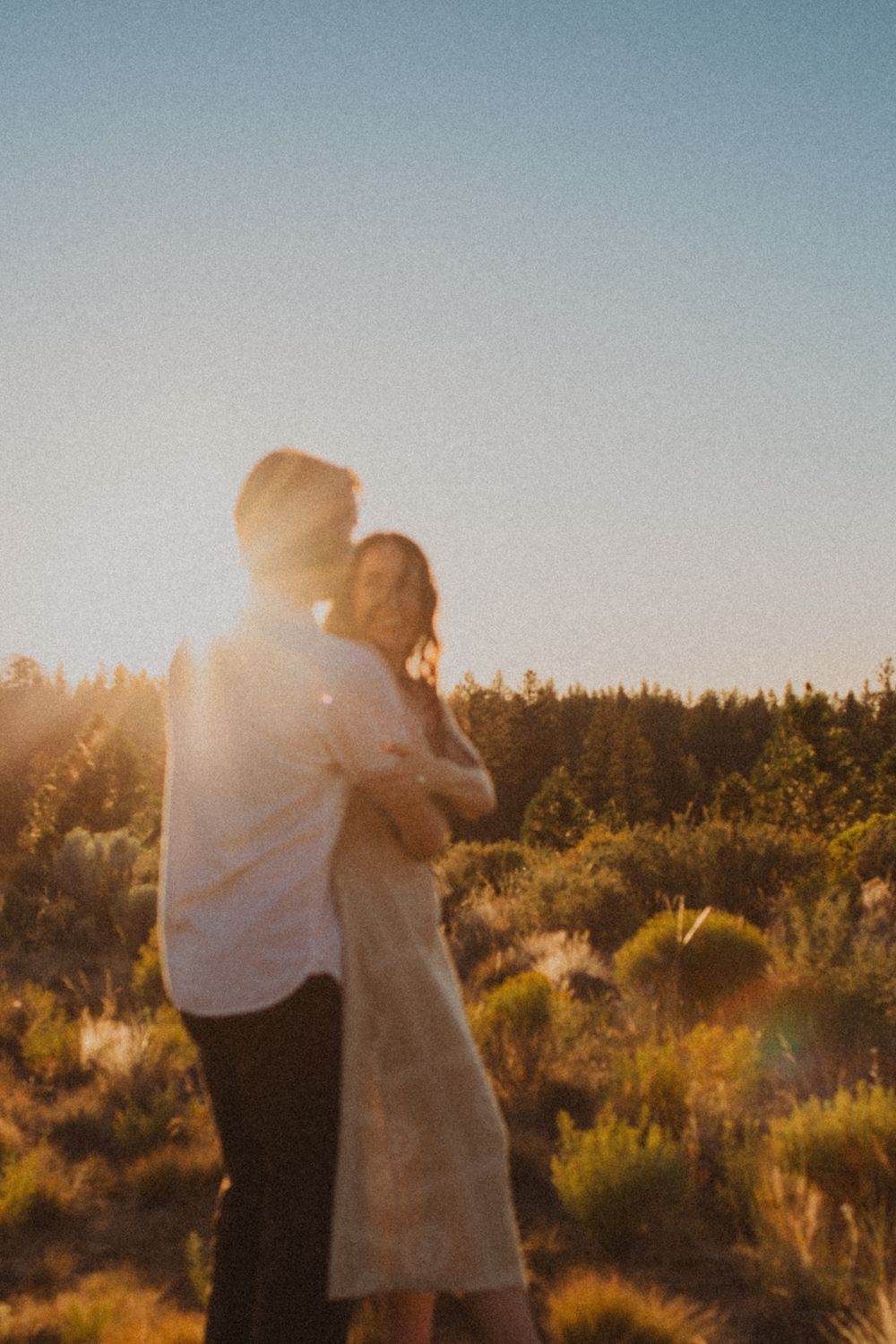 A couple embraces in a desert landscape at golden hour with sunlight streaming through the trees.