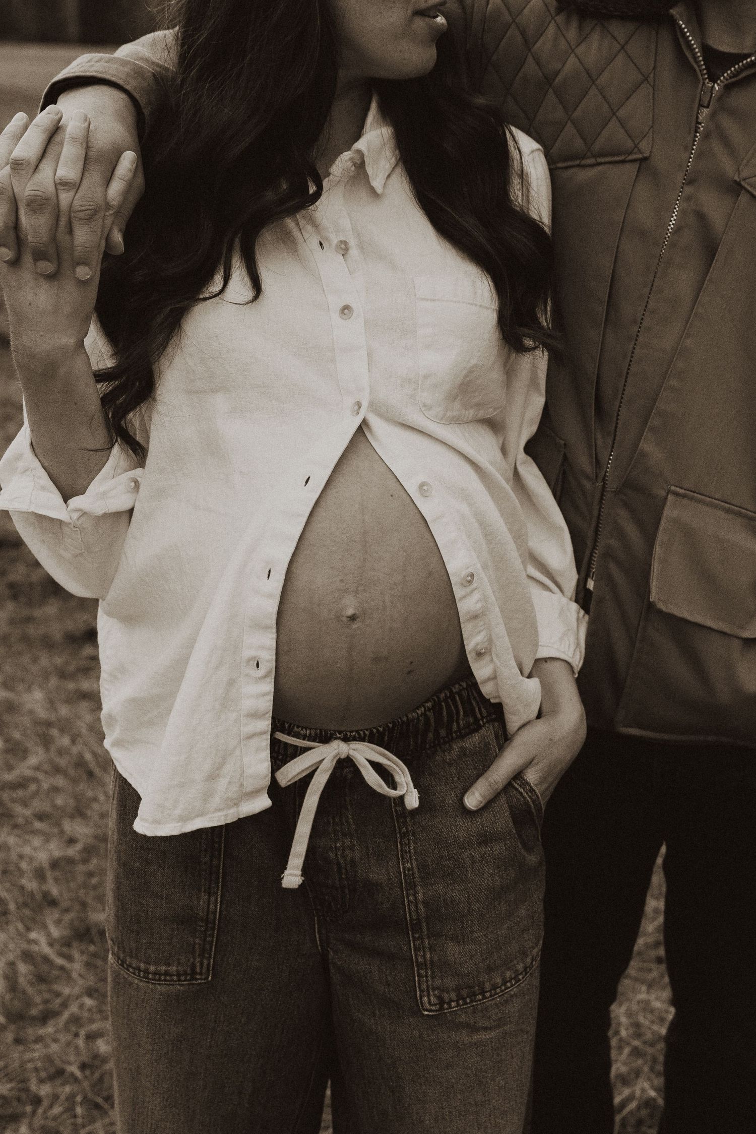 Sepia close-up of pregnant couple standing together, woman cradling bare baby bump in open white shirt.