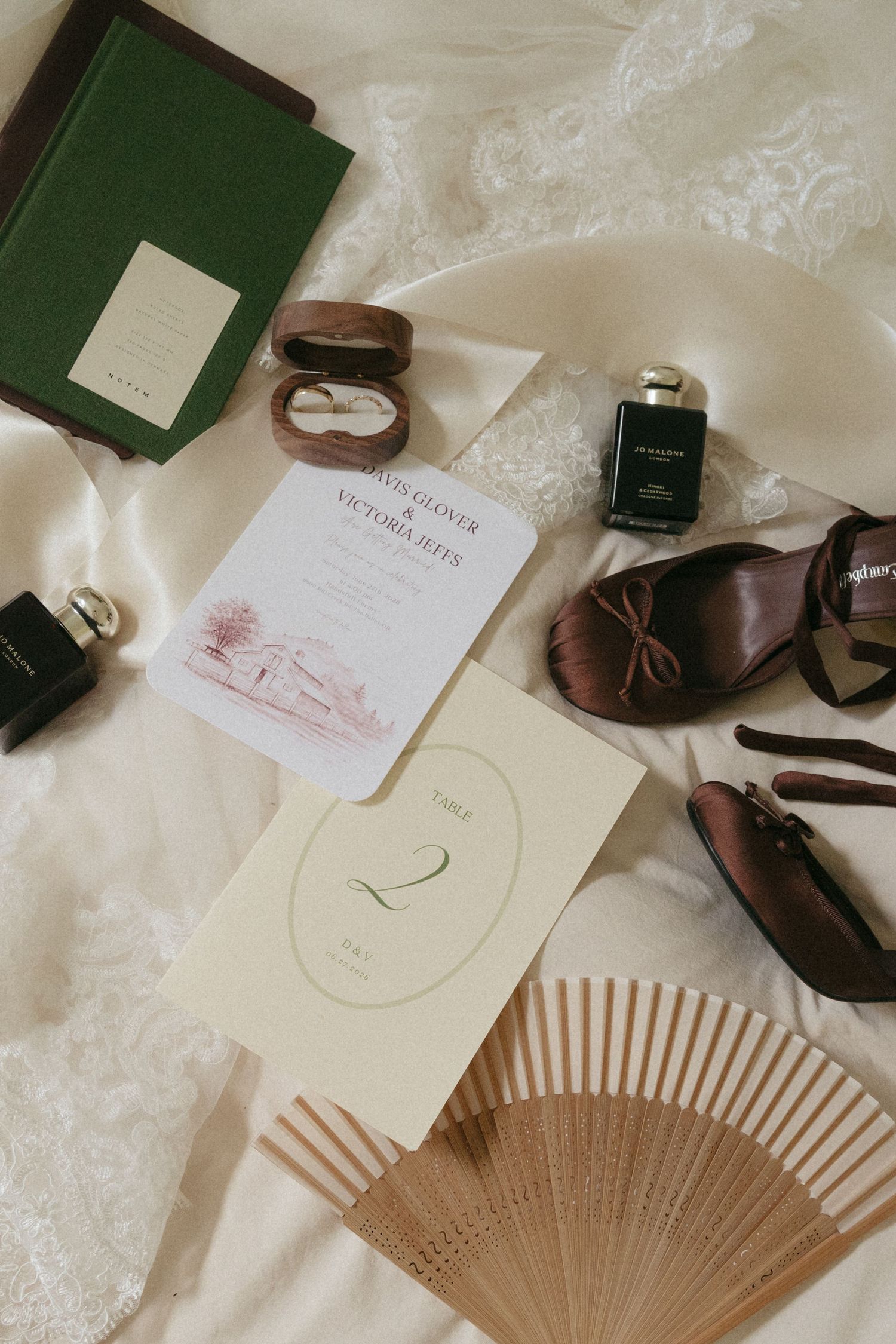 Wedding flat lay with invitation, table number, fan, brown heels, perfume, ring box, and green book on white fabric.