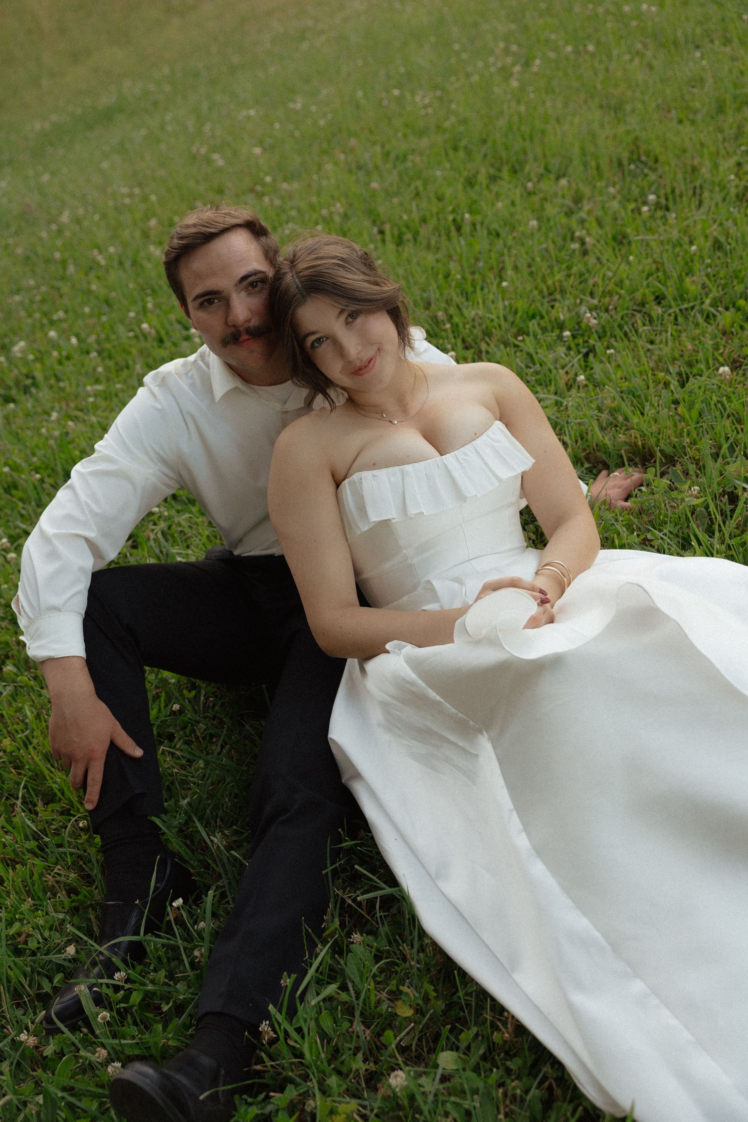 Bride and groom sitting together on green grass, smiling at camera during outdoor wedding photoshoot.