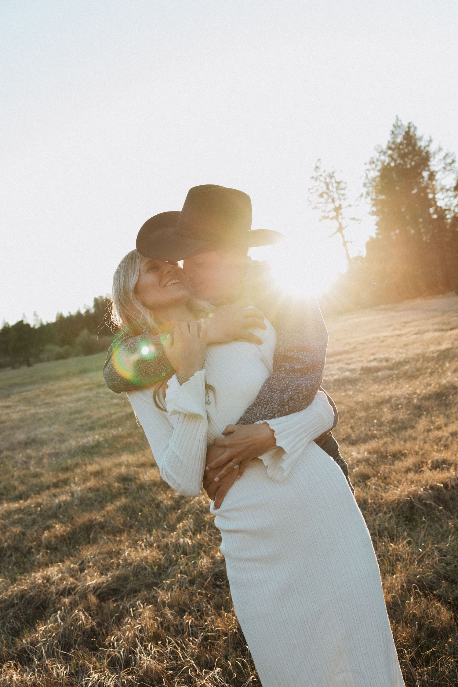 Couple embracing and kissing in a sunlit golden field at sunset, woman in white dress and man in black cowboy hat.