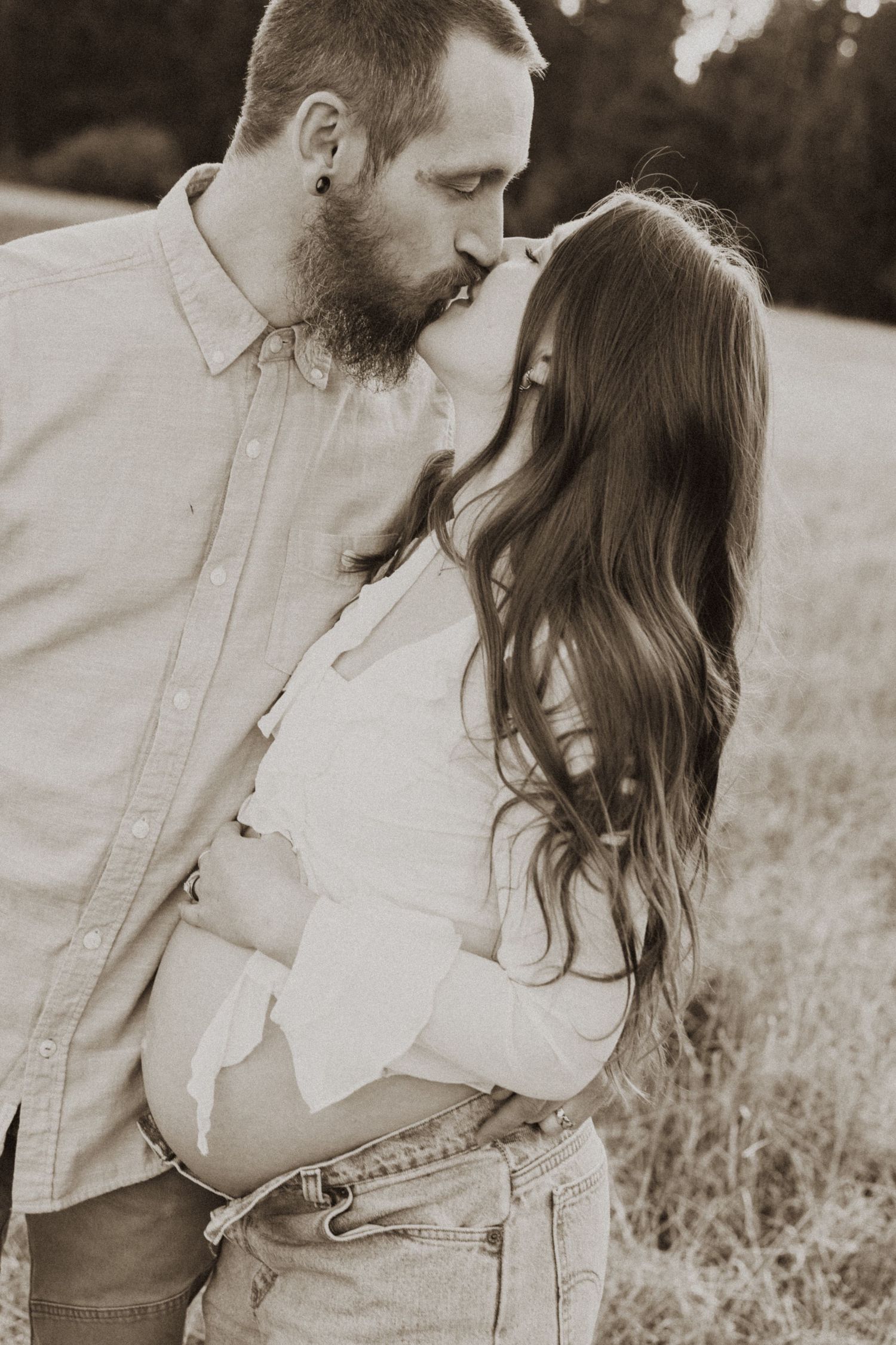 Sepia-toned maternity photo of a bearded man kissing a pregnant woman outdoors in a field.