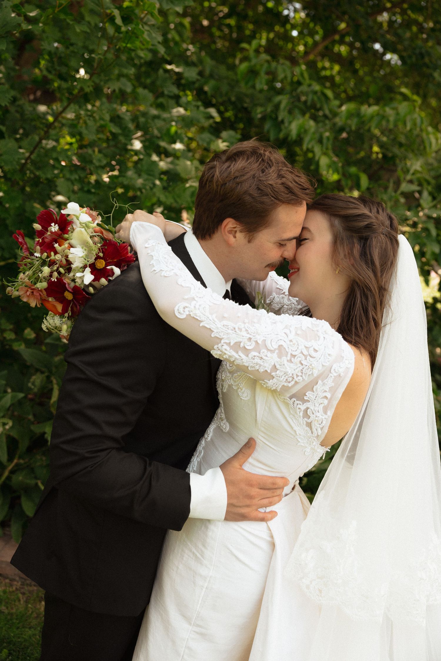Bride and groom embrace tenderly, touching foreheads in a romantic outdoor wedding portrait surrounded by lush greenery.