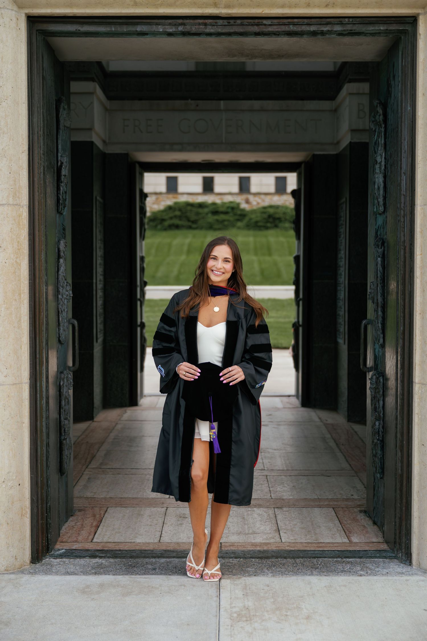 Young woman in graduation gown smiling in doorway of academic building with green lawn behind her.