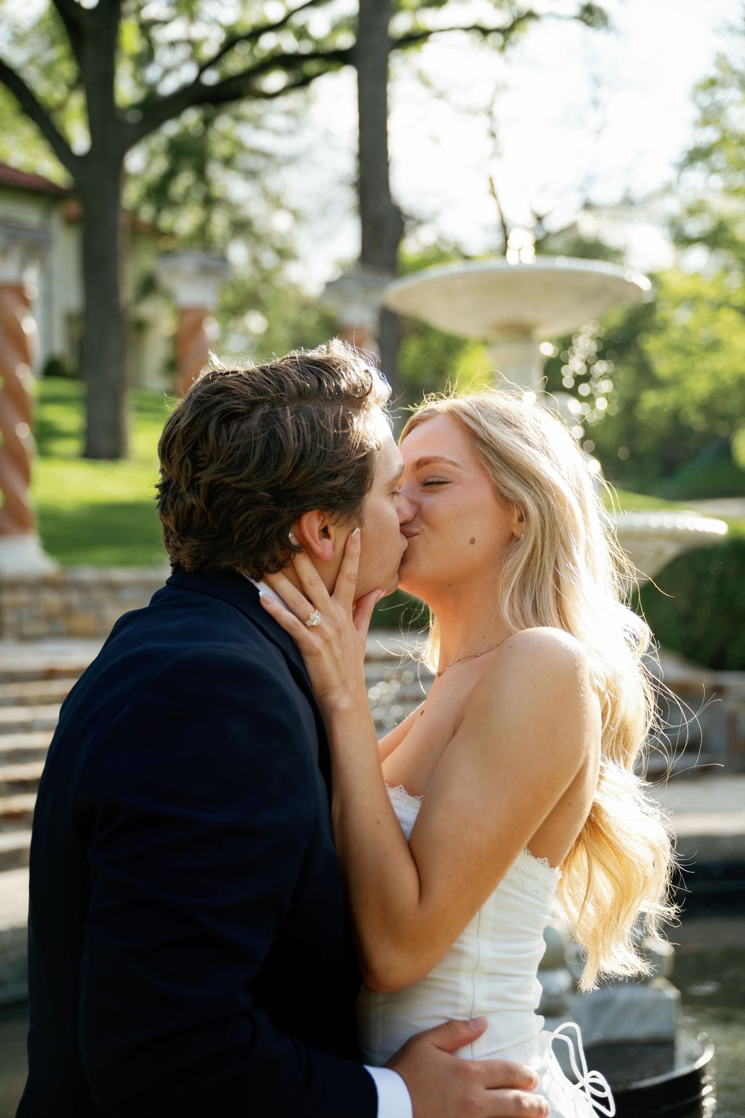 A bride and groom share a romantic kiss outdoors near a fountain, surrounded by lush greenery on their wedding day.
