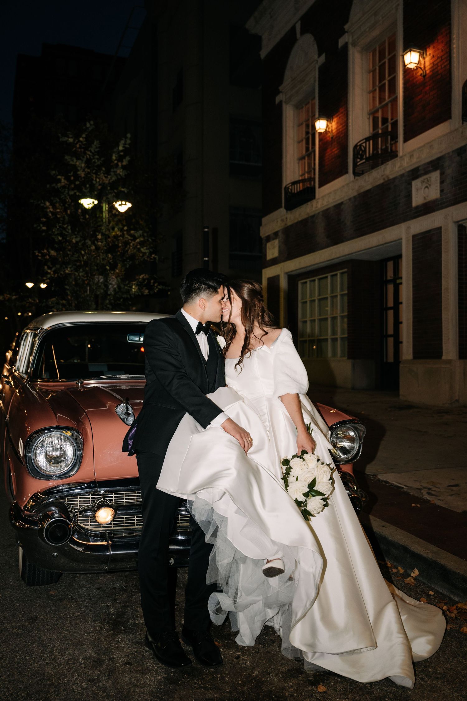 A couple in wedding attire poses dramatically at night with vintage cars in the background.