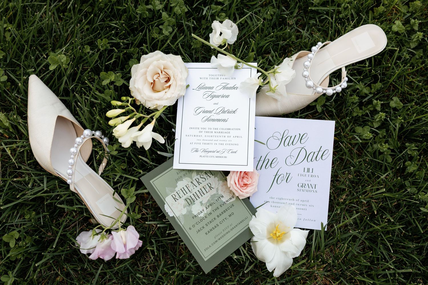Wedding stationery suite with save the date cards, invitations, and pearl-adorned bridal heels on green grass with flowers.