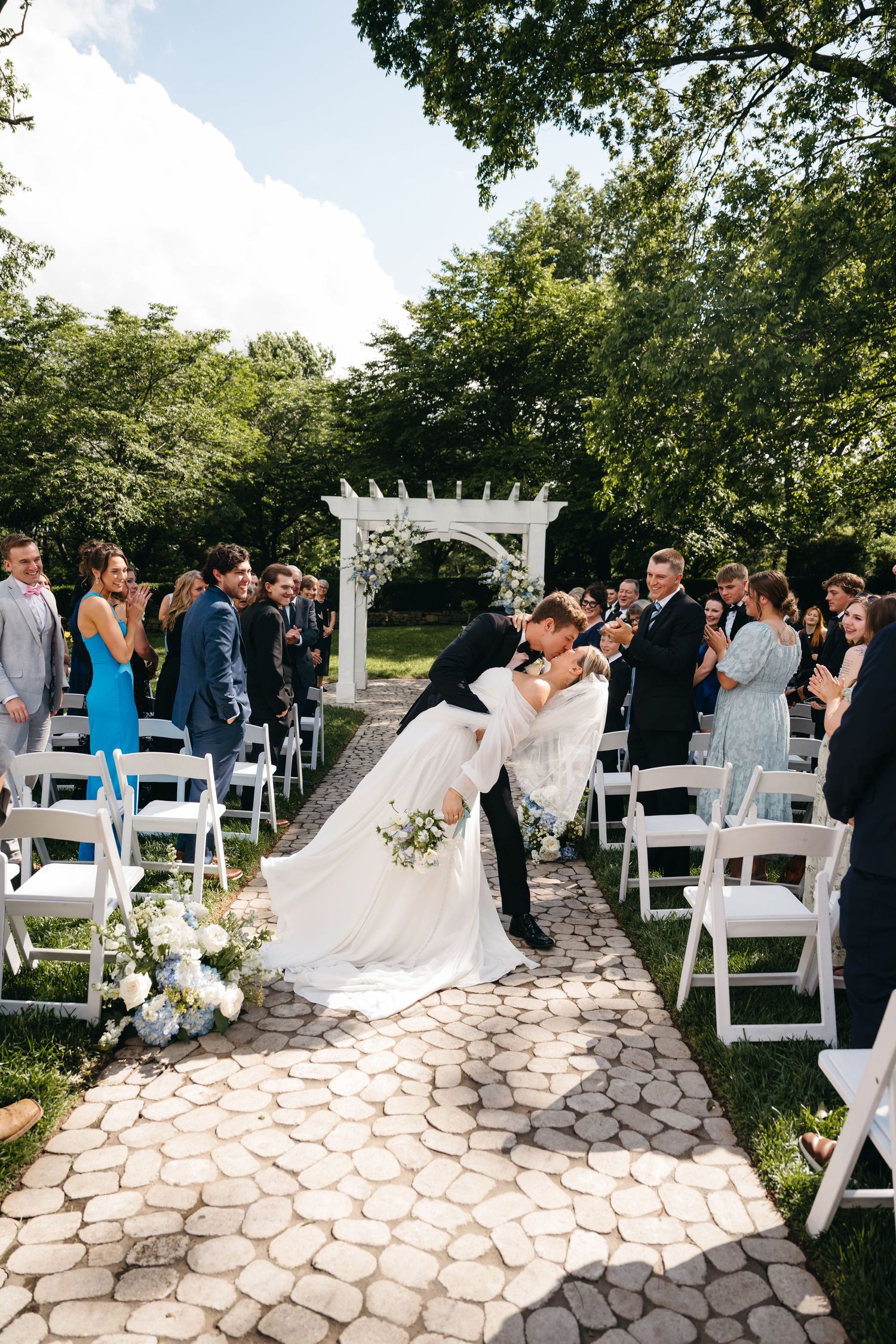 A romantic outdoor wedding ceremony takes place on a stone paved pathway surrounded by white chairs and greenery.