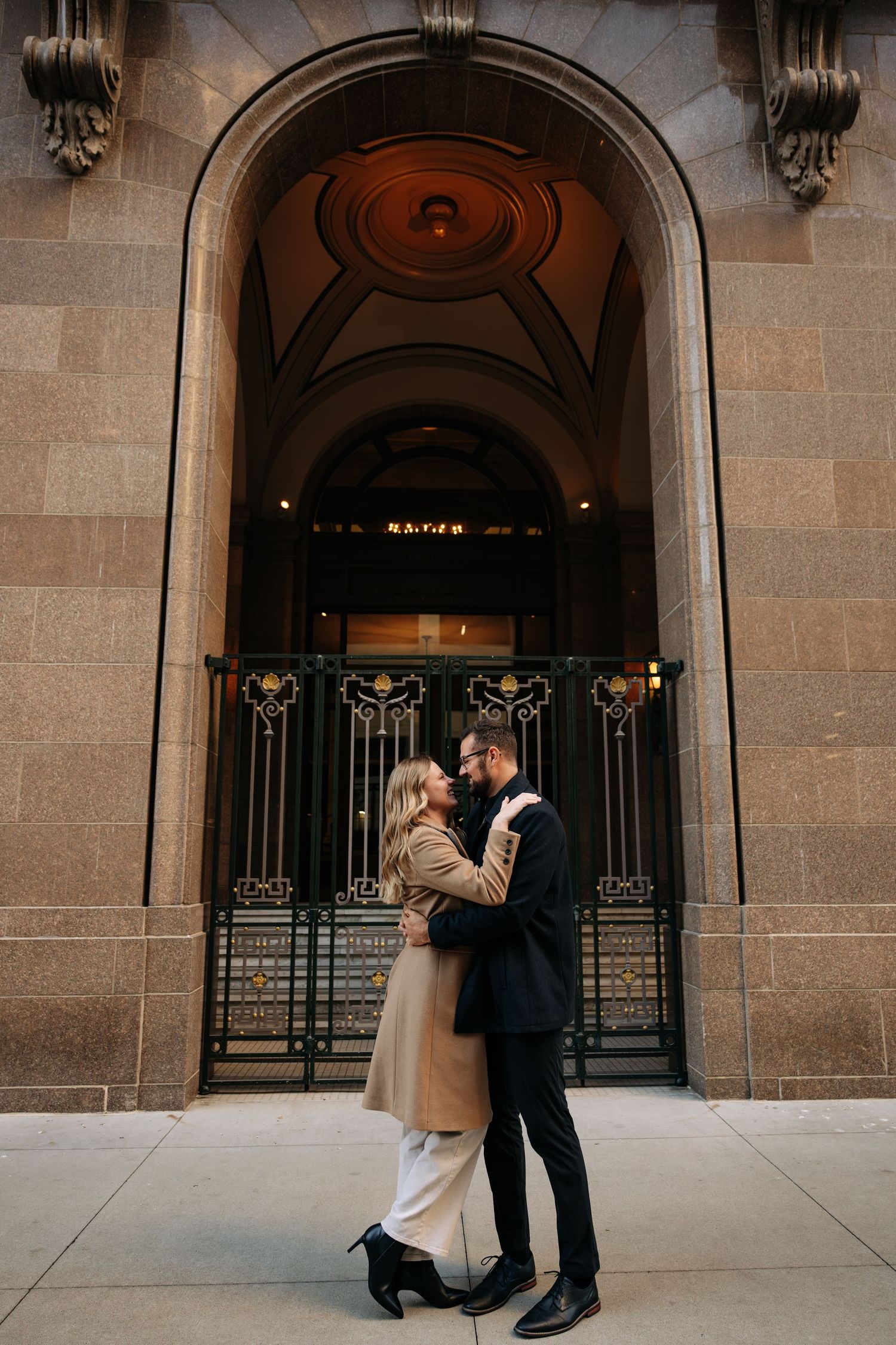 A couple shares an intimate moment in front of an ornate stone building entrance.
