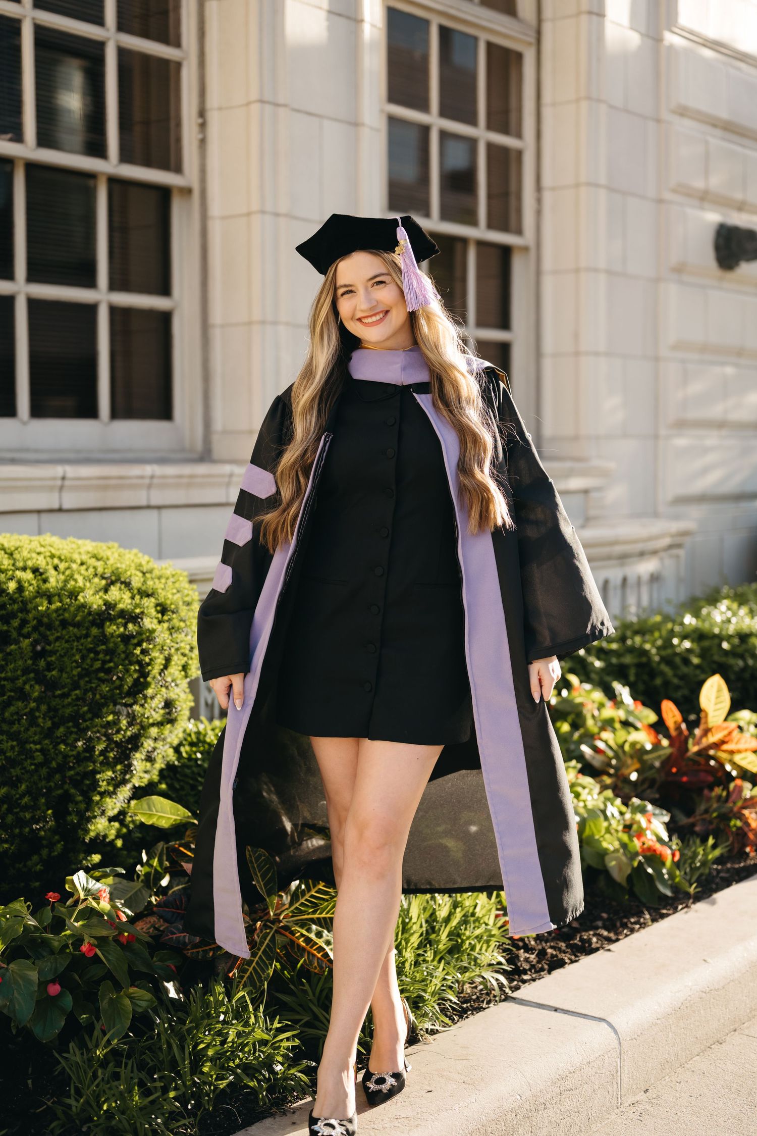 Graduate in black cap and gown with purple stole poses in front of university building with landscaping.