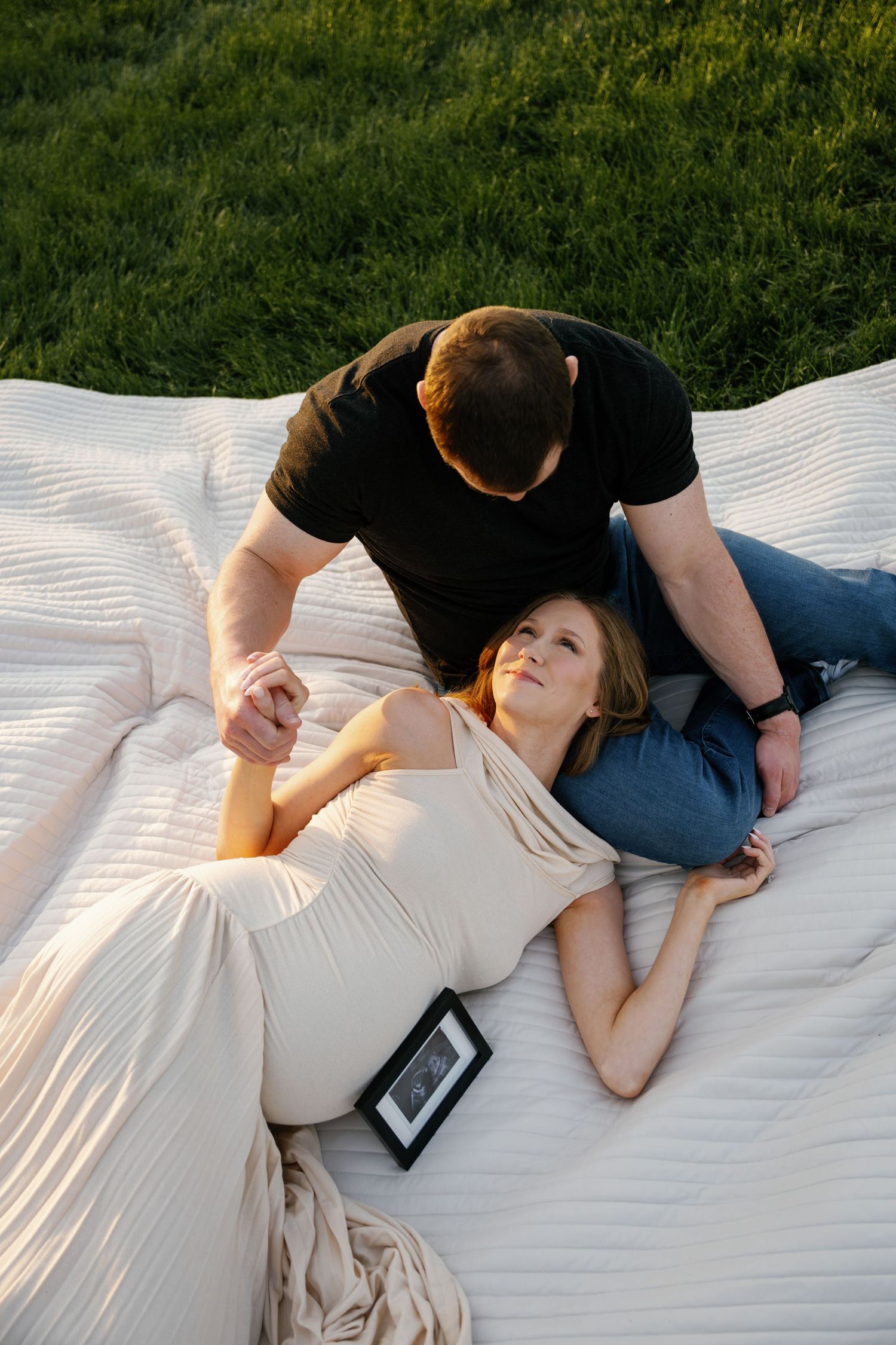 Pregnant woman in beige dress lying on white blanket outdoors, holding hands with man in black shirt beside her.