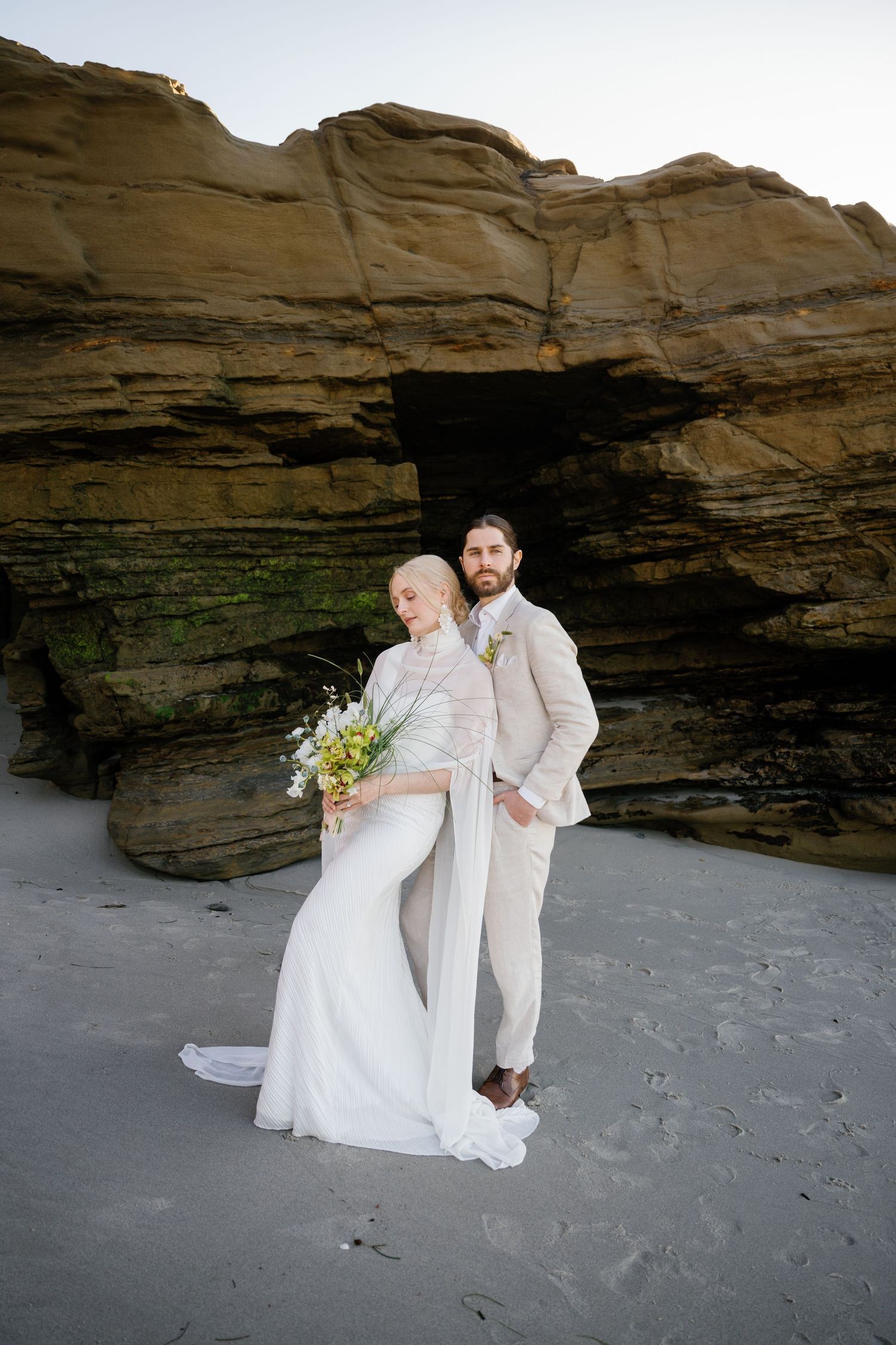 Wedding couple poses under dramatic rock formations on sandy beach.