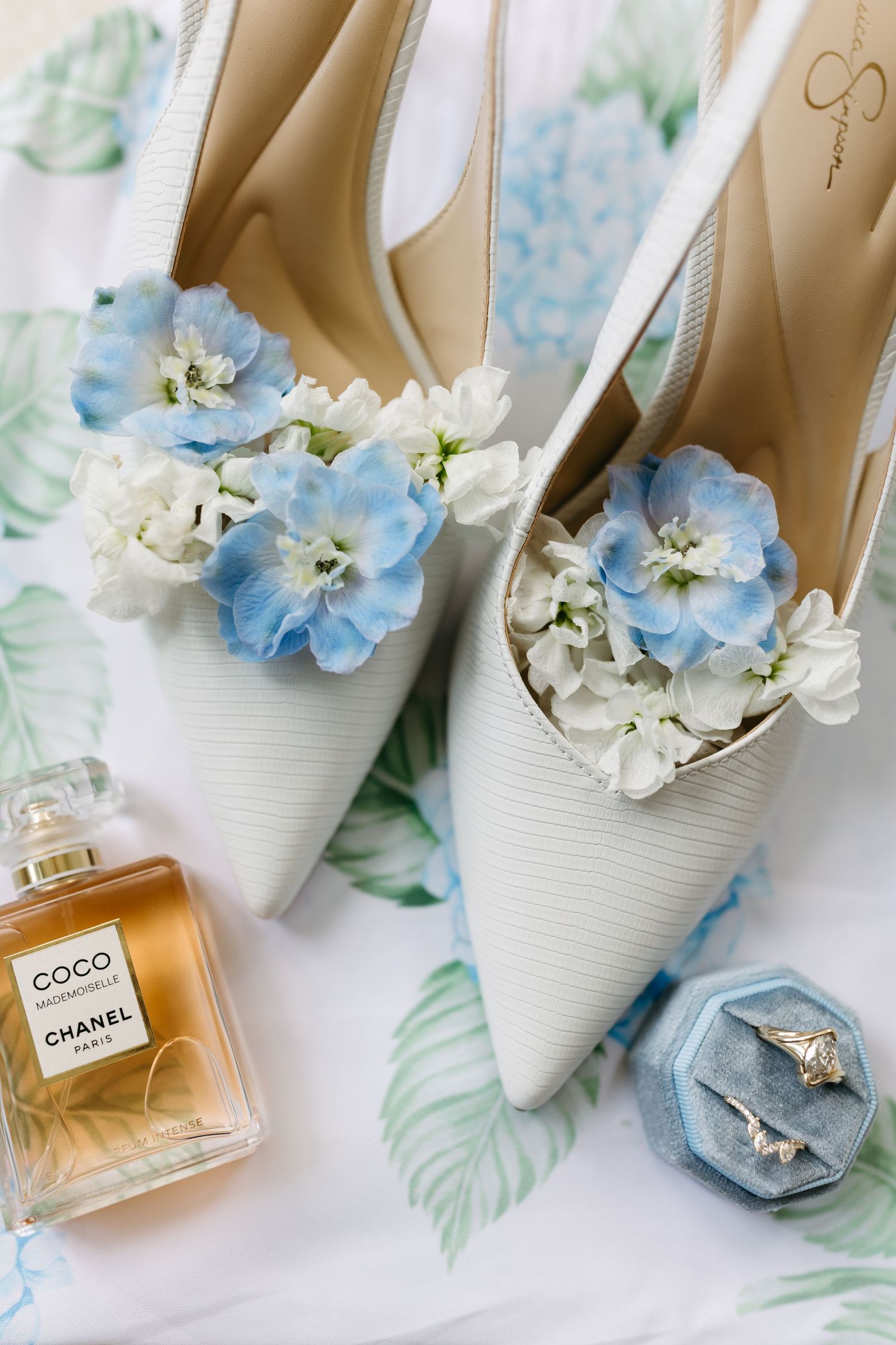 Wedding accessories including white shoes, blue flowers, and Chanel perfume bottles arranged on floral fabric.