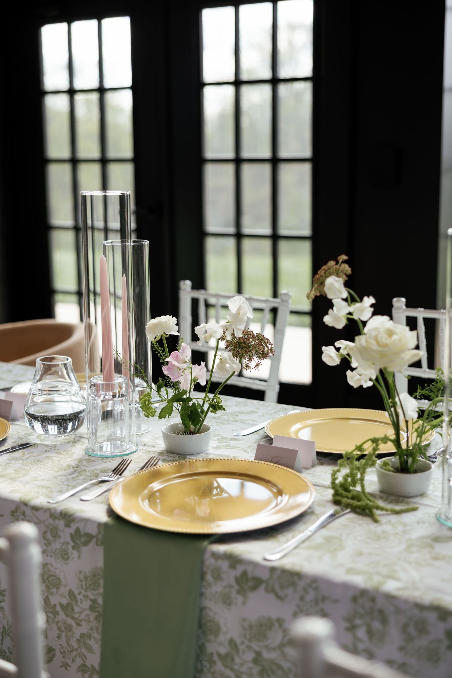 Elegant wedding table setting with gold charger plates, white floral centerpieces, and sage green tablecloth.