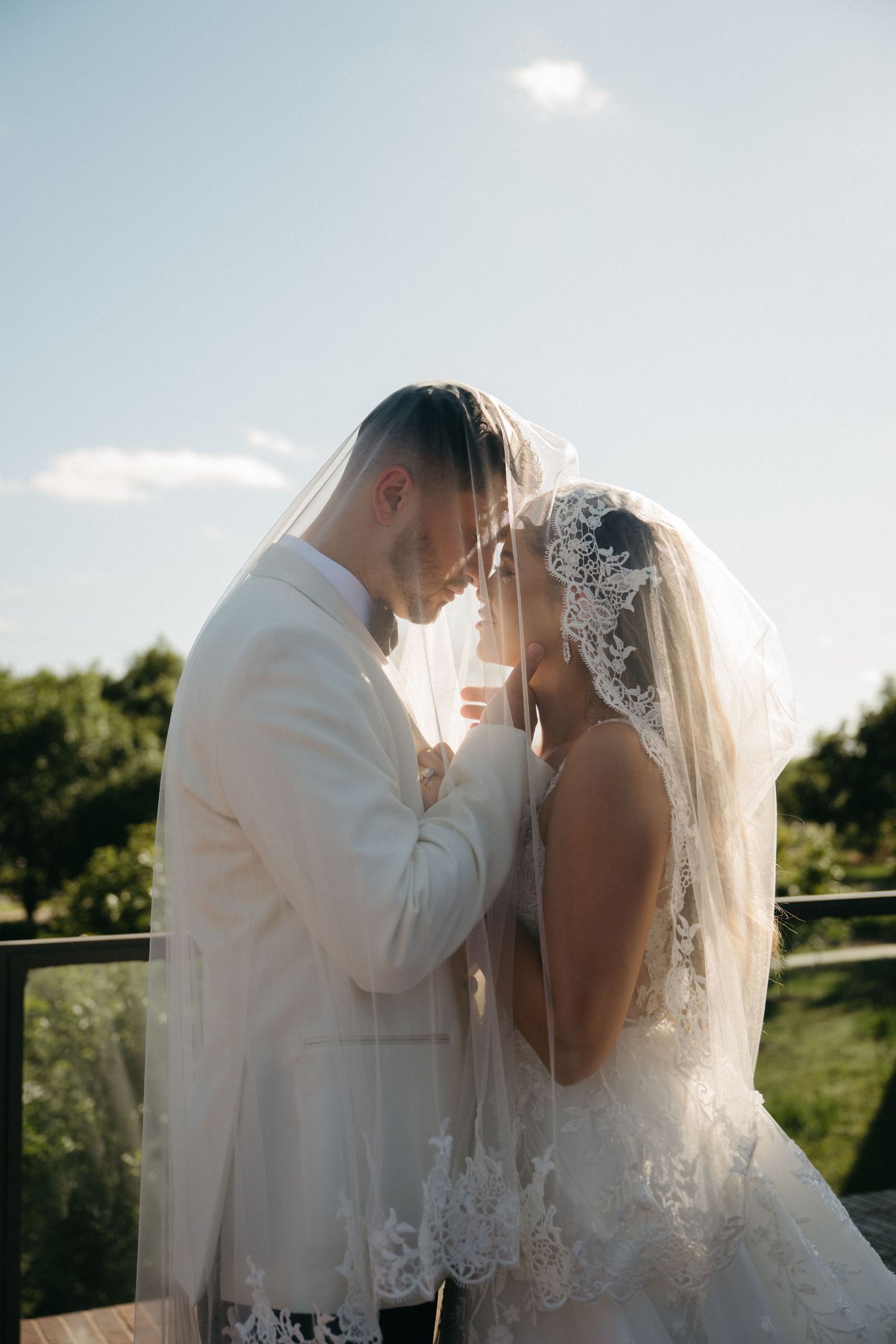 A romantic wedding couple embraces outdoors during golden hour with a scenic backdrop of trees and sunlight.