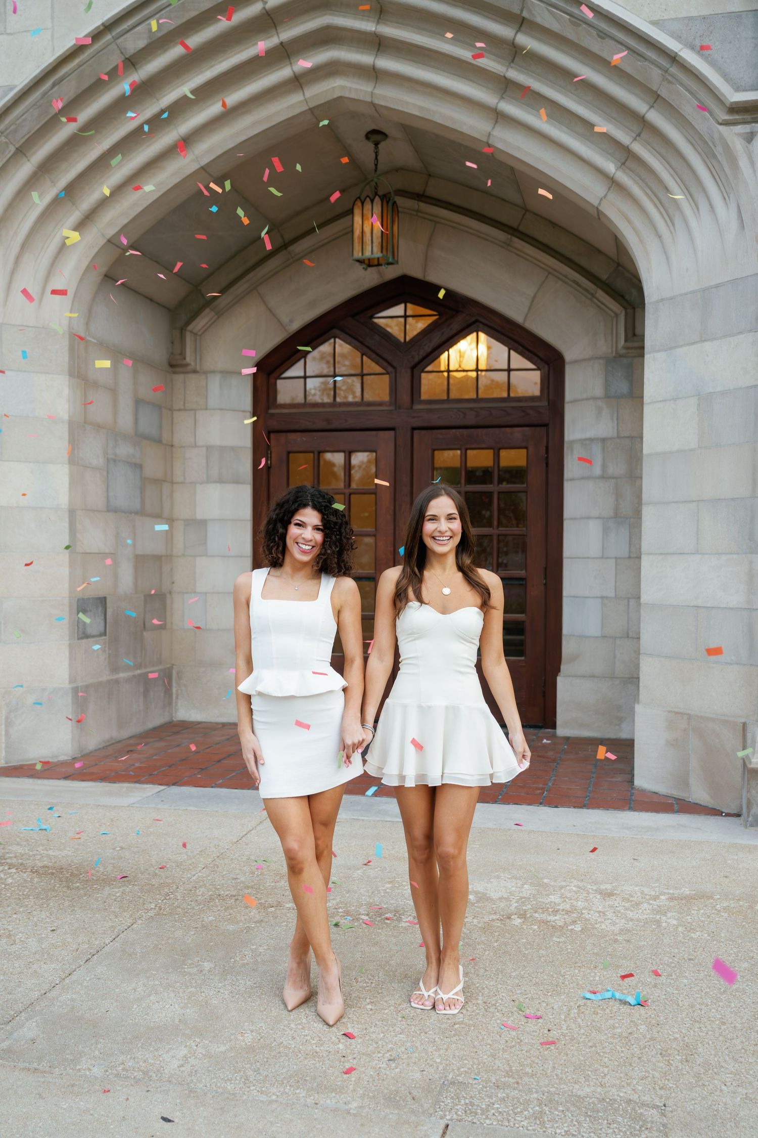 Two women in white dresses hold hands under a stone archway as colorful confetti falls around them.