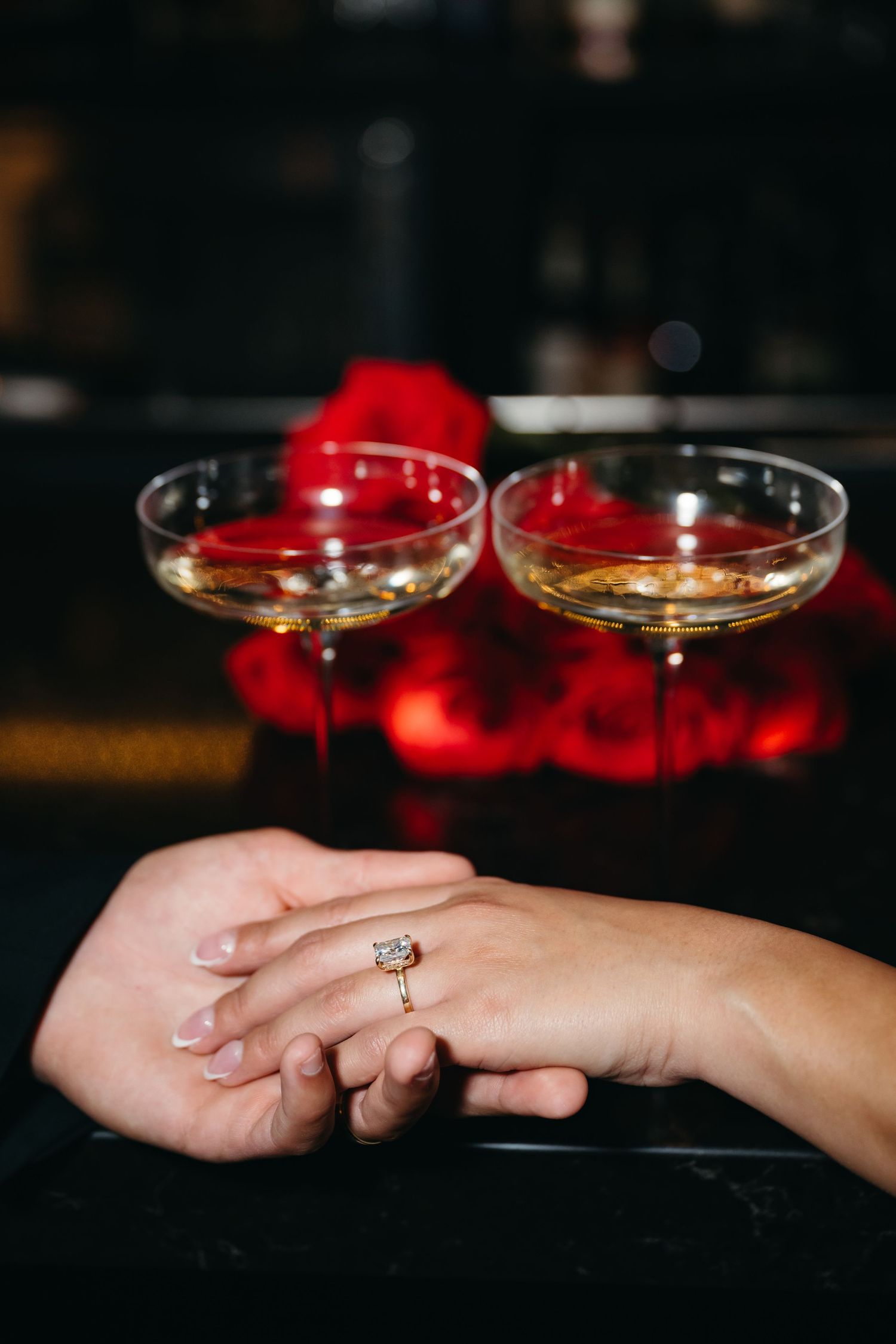Close-up of hands holding champagne glasses with red rose petals in the background.