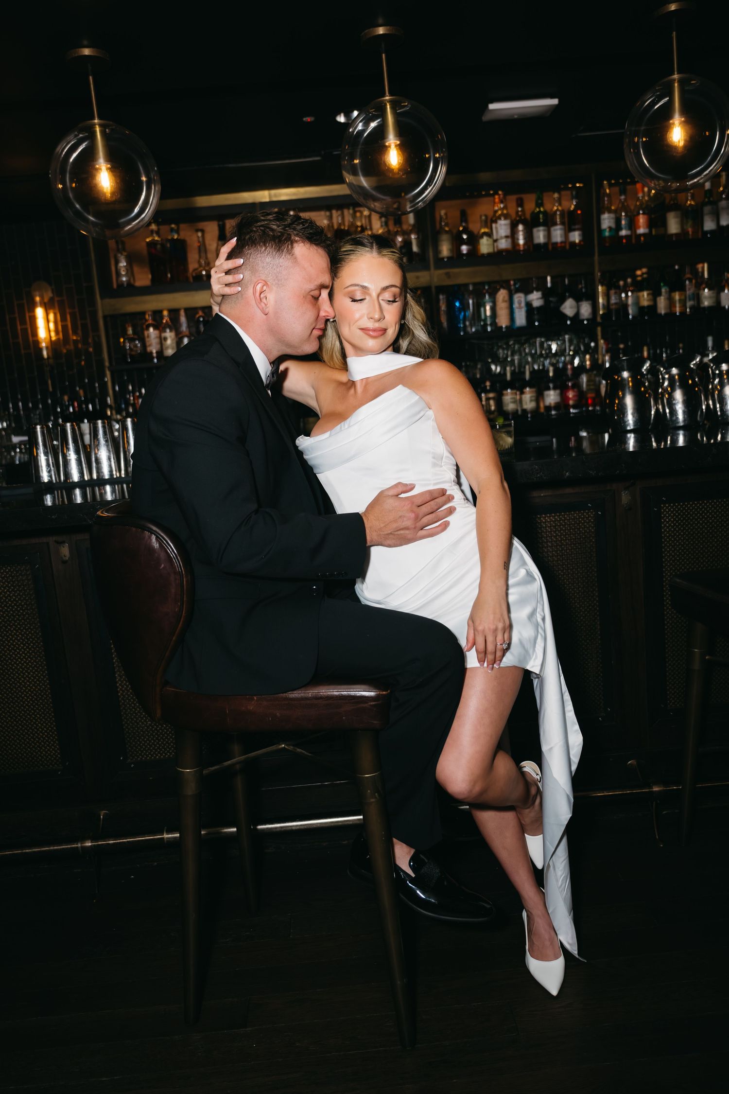 Elegant bride and groom sharing an intimate moment at a dark bar, wearing white dress and black tuxedo.