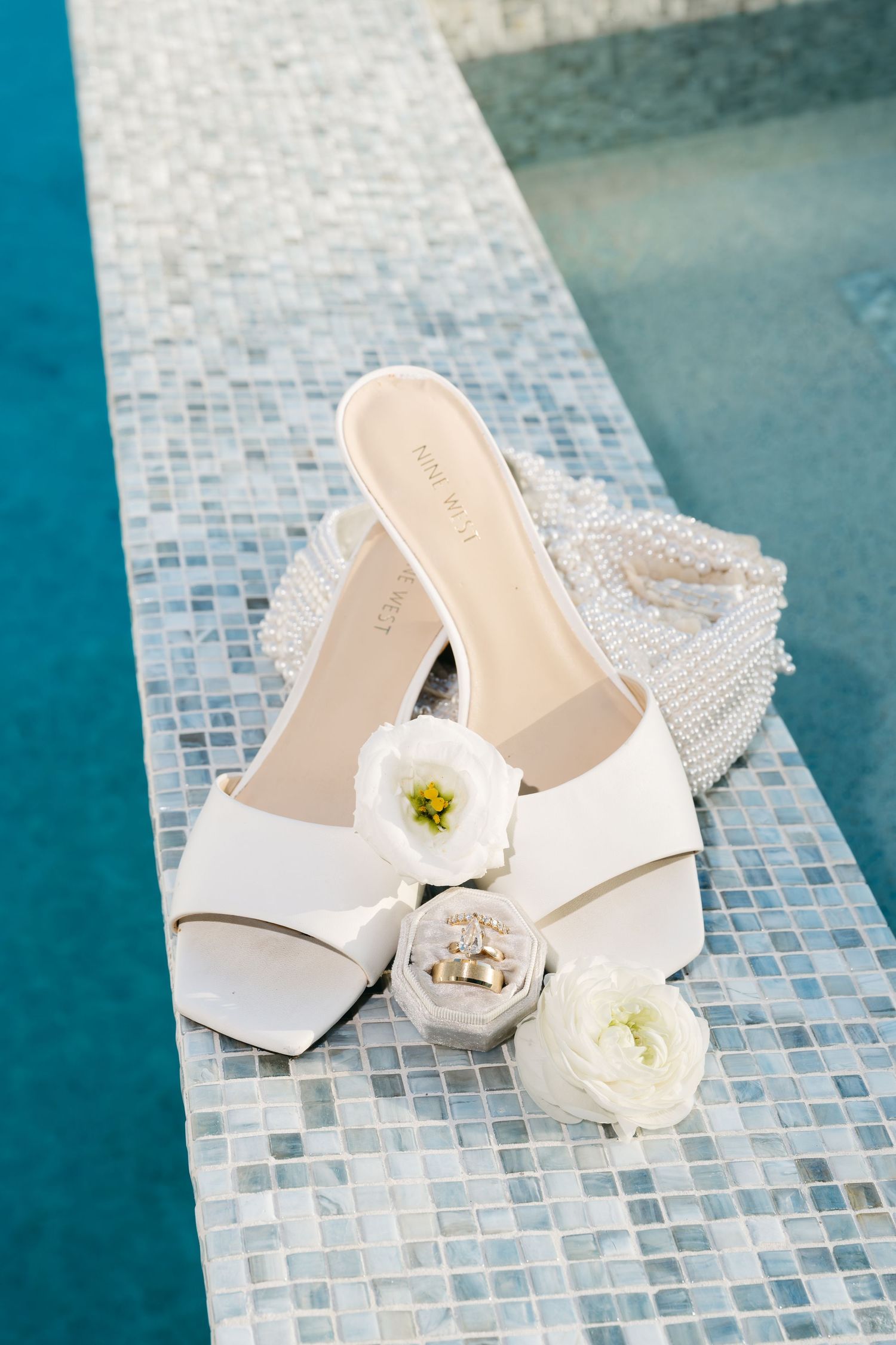 White bridal heels with floral detail, pearl clutch, and wedding rings on mosaic tile poolside.