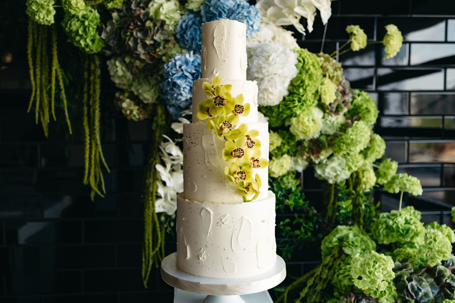 Elegant white wedding cake decorated with delicate sugar flowers and greenery against a dark floral backdrop.