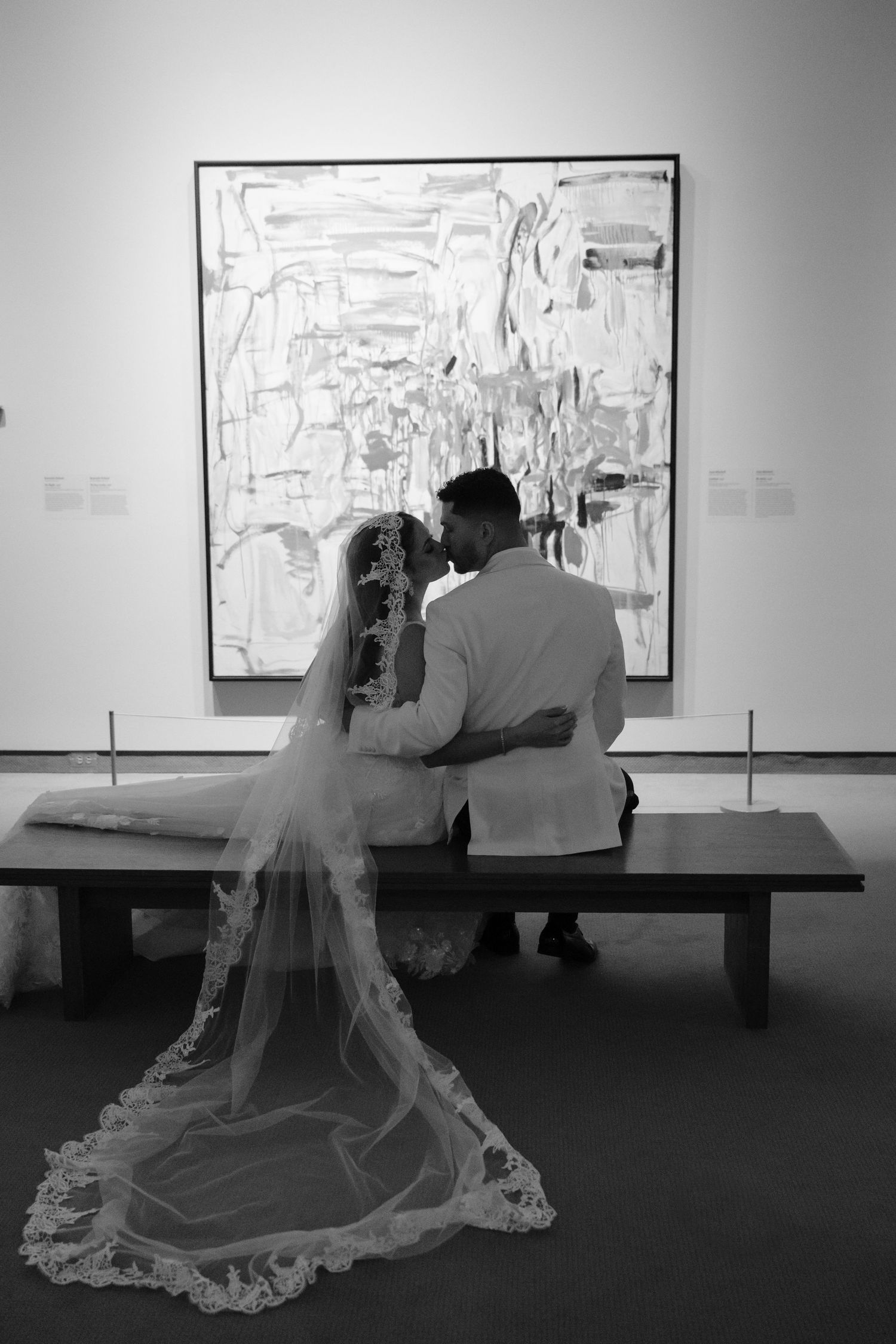 A couple in wedding attire sits on a bench viewing abstract artwork in a gallery, with a flowing lace veil trailing behind.