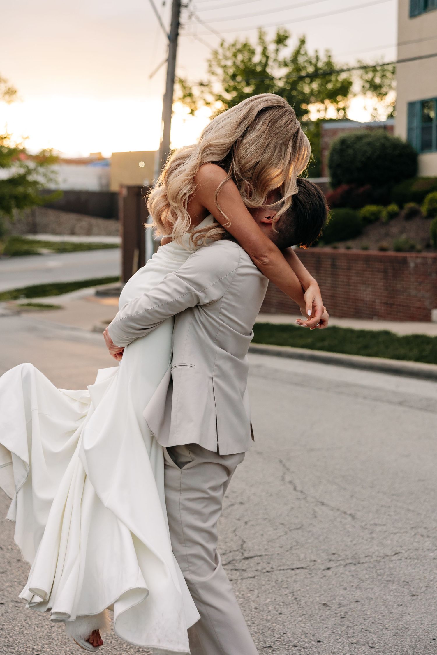 A romantic wedding couple dips and embraces on a sunlit street in formal attire.