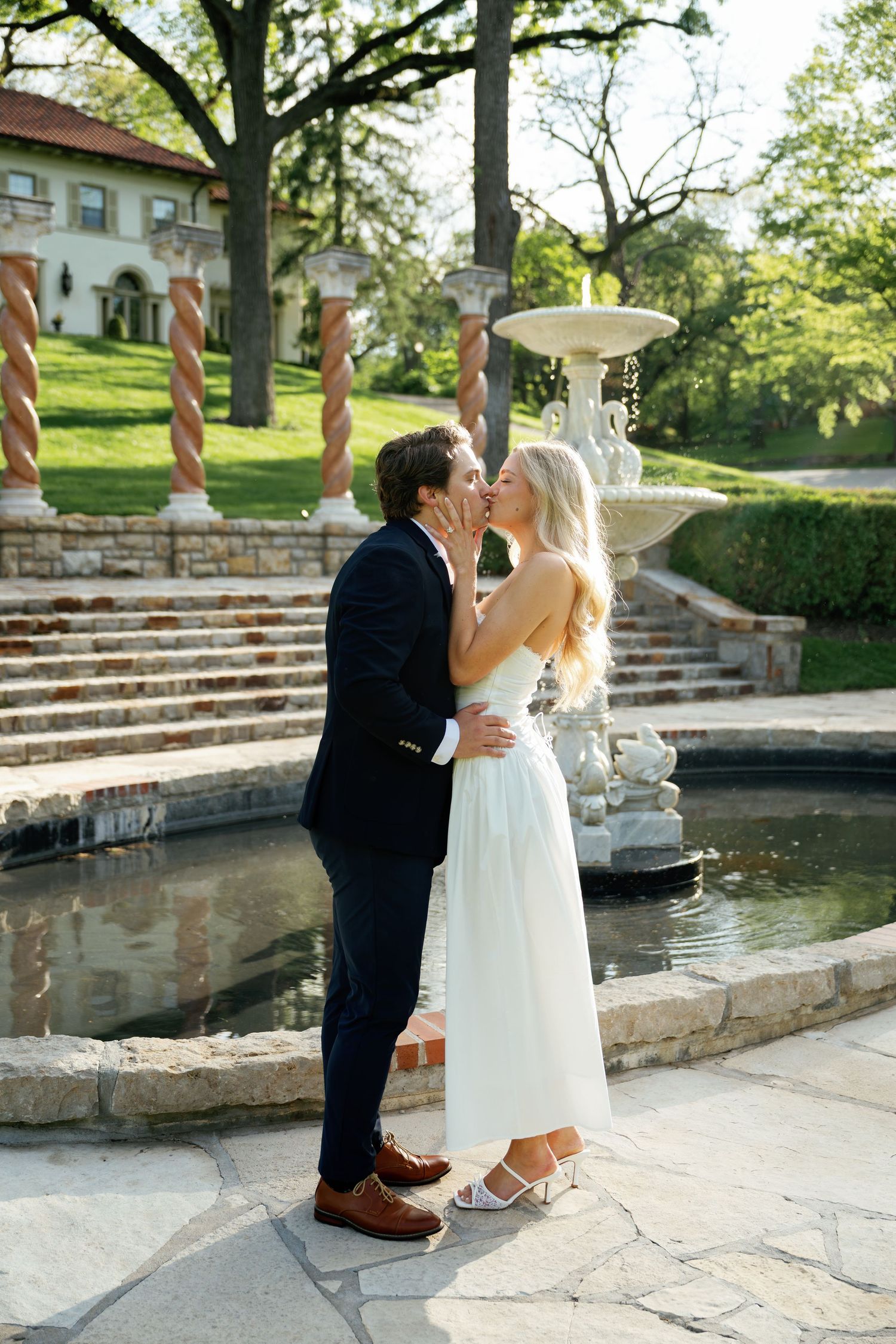 Bride and groom sharing a romantic kiss by a stone fountain in an elegant garden venue on their wedding day.