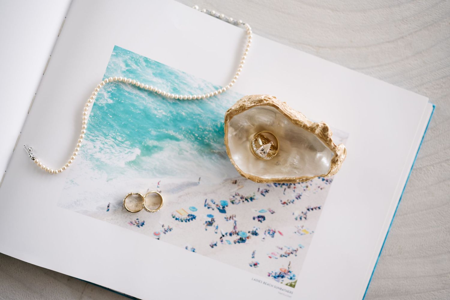 Pearl necklace and gold earrings displayed in an oyster shell on an open book with watercolor art.