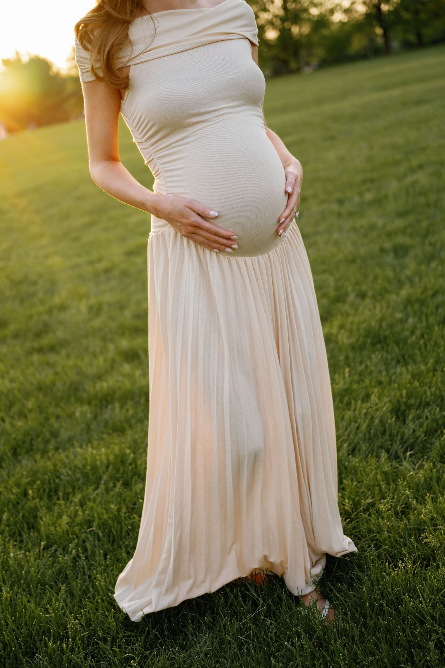 Pregnant woman in elegant cream dress cradling baby bump outdoors at golden hour sunset on green grass.