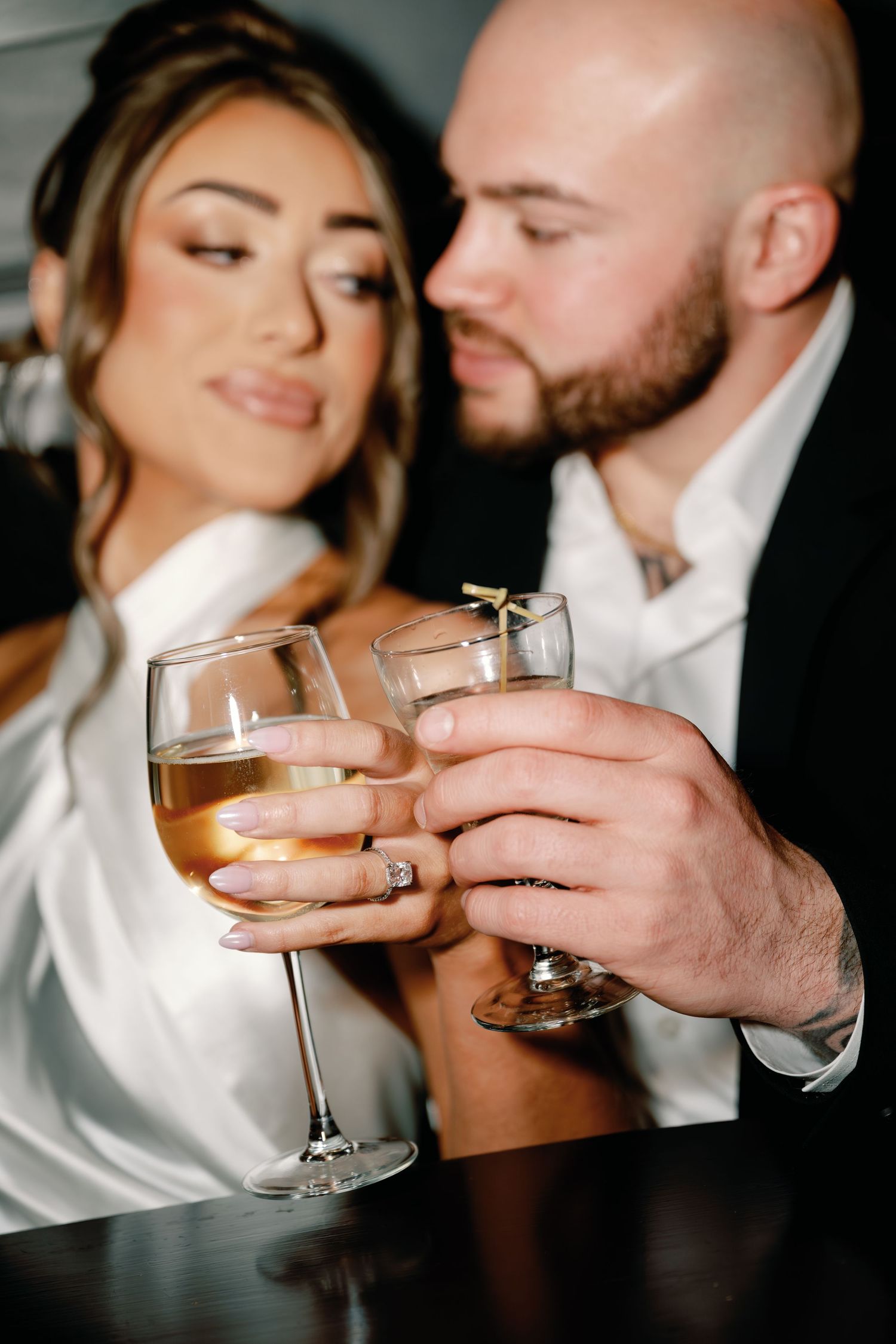 Elegant couple toasting with wine glasses, bride in white dress with diamond ring, groom in black suit at romantic event.