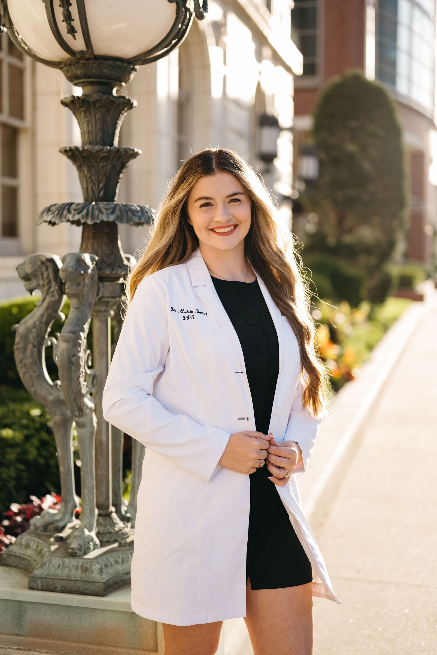 Graduate wearing white lab coat poses confidently outdoors near ornate light fixtures.