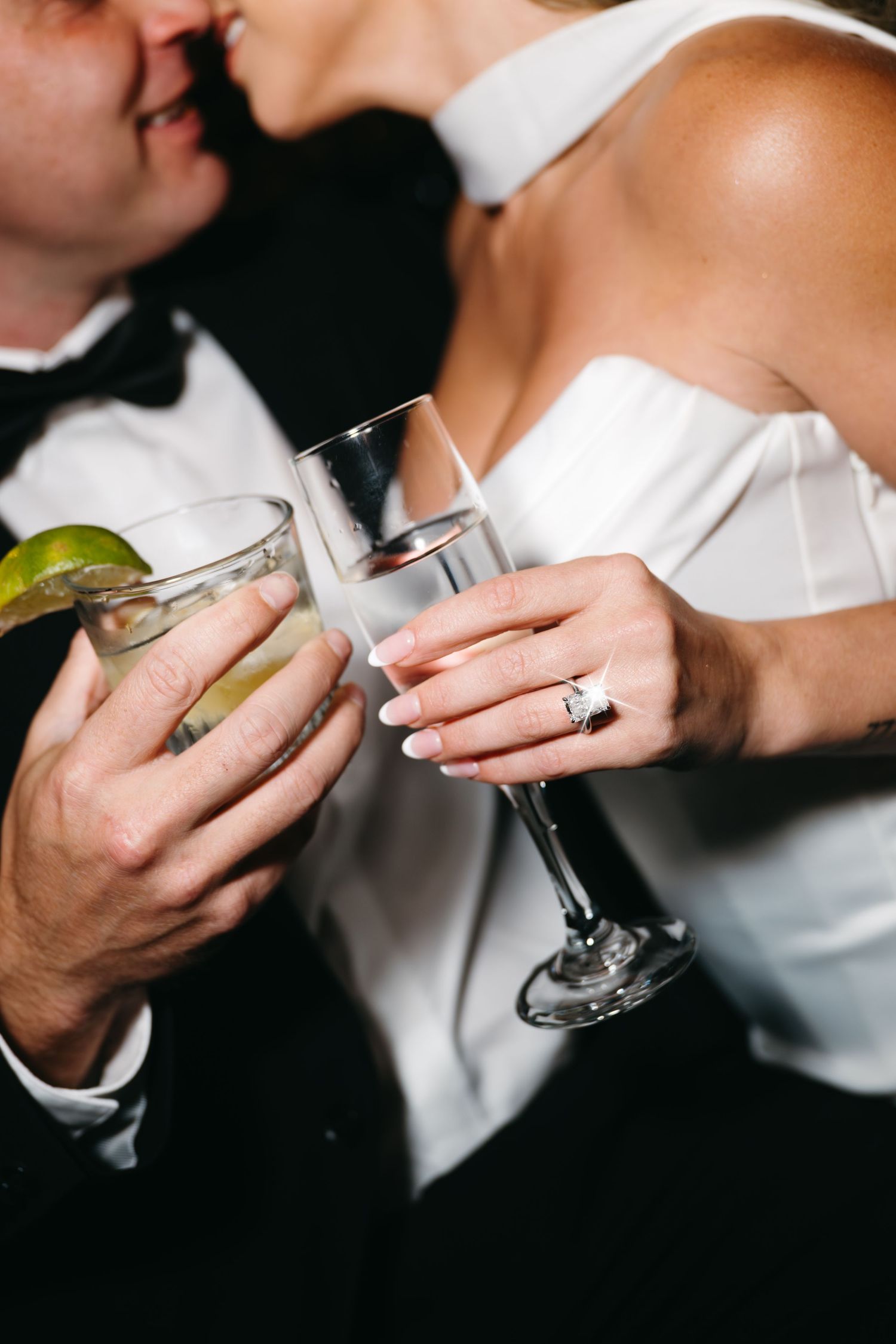 Bride and groom clinking glasses at wedding reception, woman wearing diamond engagement ring, man in tuxedo.