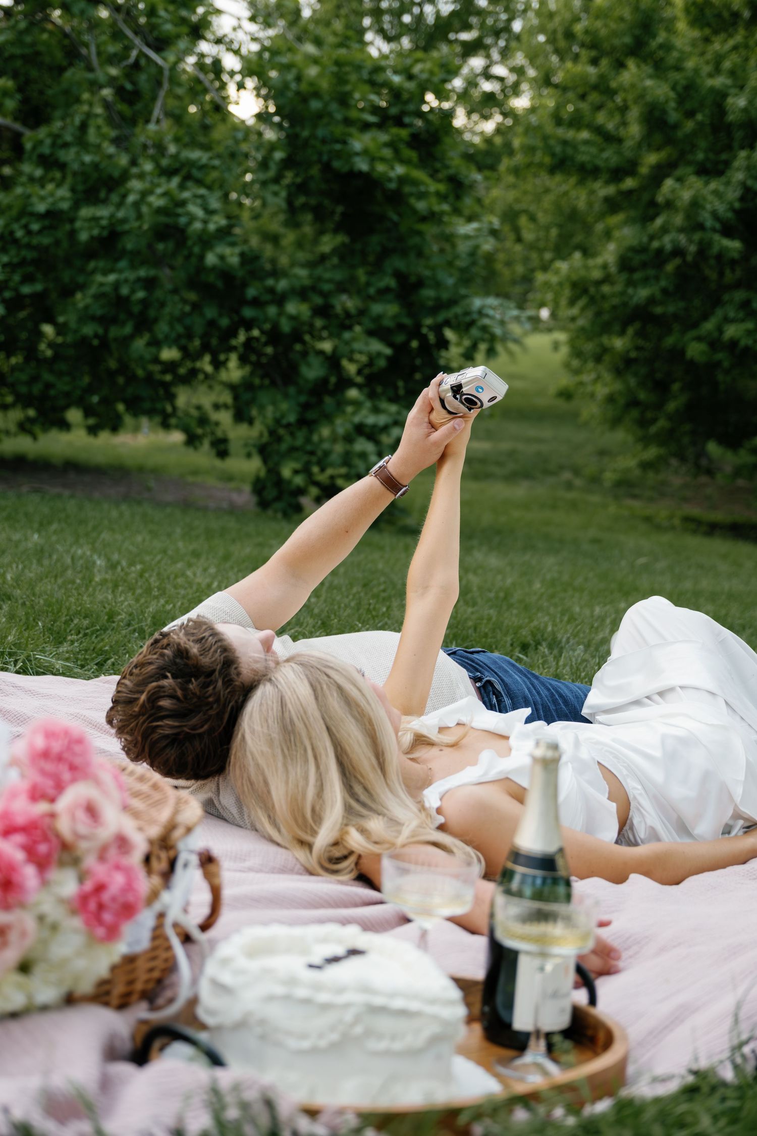 Couple lying on a pink blanket at a romantic outdoor picnic with champagne, flowers, and a cake in a lush green park.