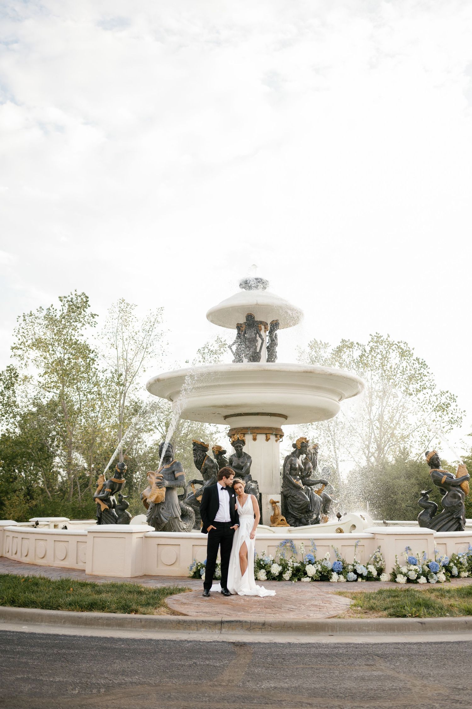 Bride and groom share a romantic kiss in front of an elegant white fountain with bronze statues at their wedding.