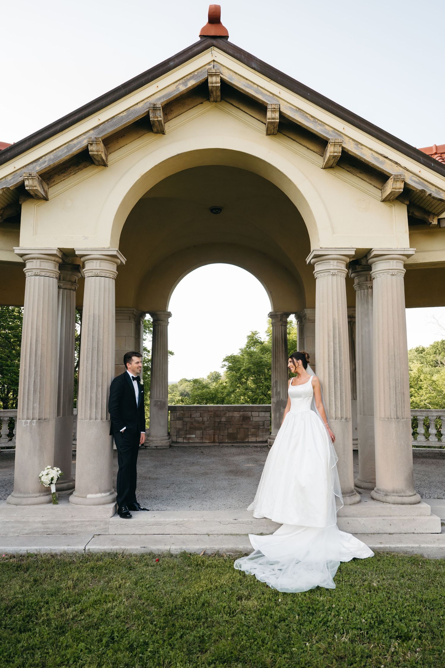 Wedding couple stands apart under elegant stone archways in a garden setting during their first look photo session.